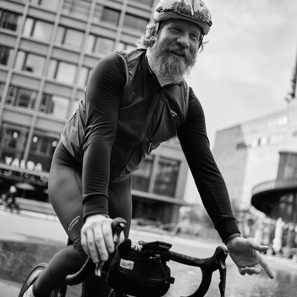 Model wearing black cycling jersey and bib shorts in urban setting, leaning over bike handlebars