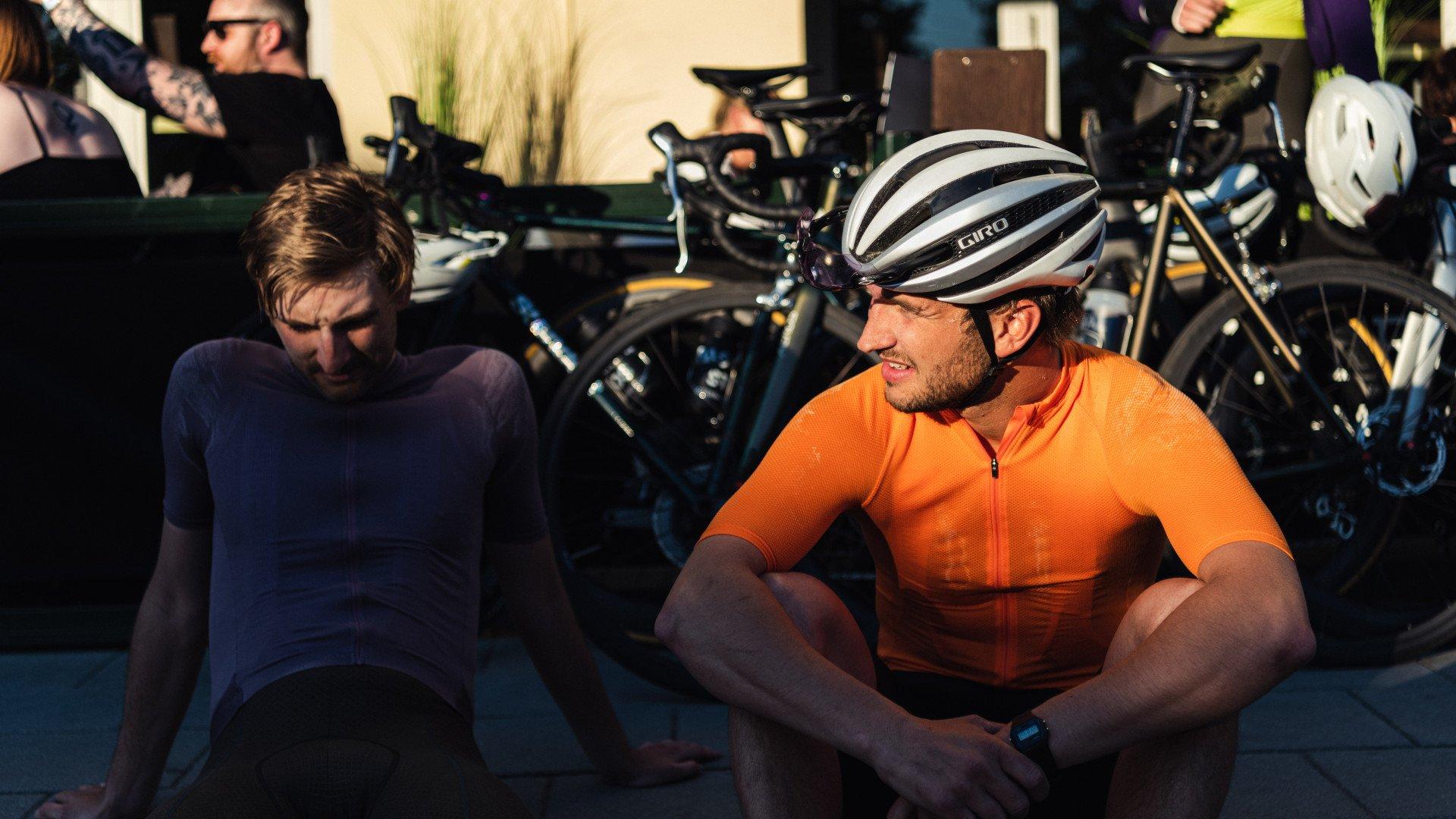 Two cyclists in orange and navy jerseys resting beside bikes in golden hour light