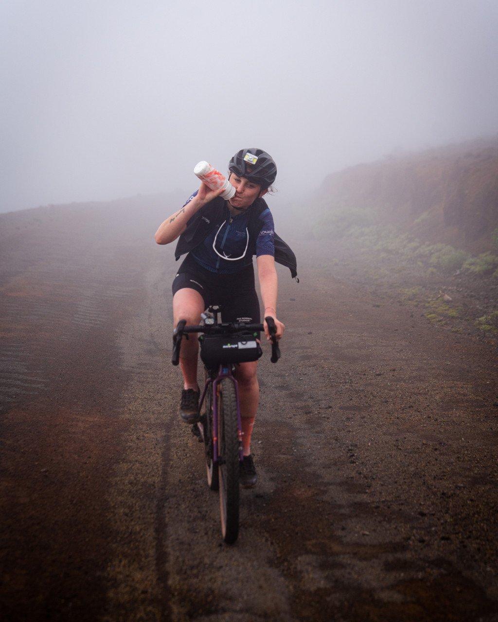 Cyclist drinking from water bottle on misty gravel road wearing dark jersey and shorts