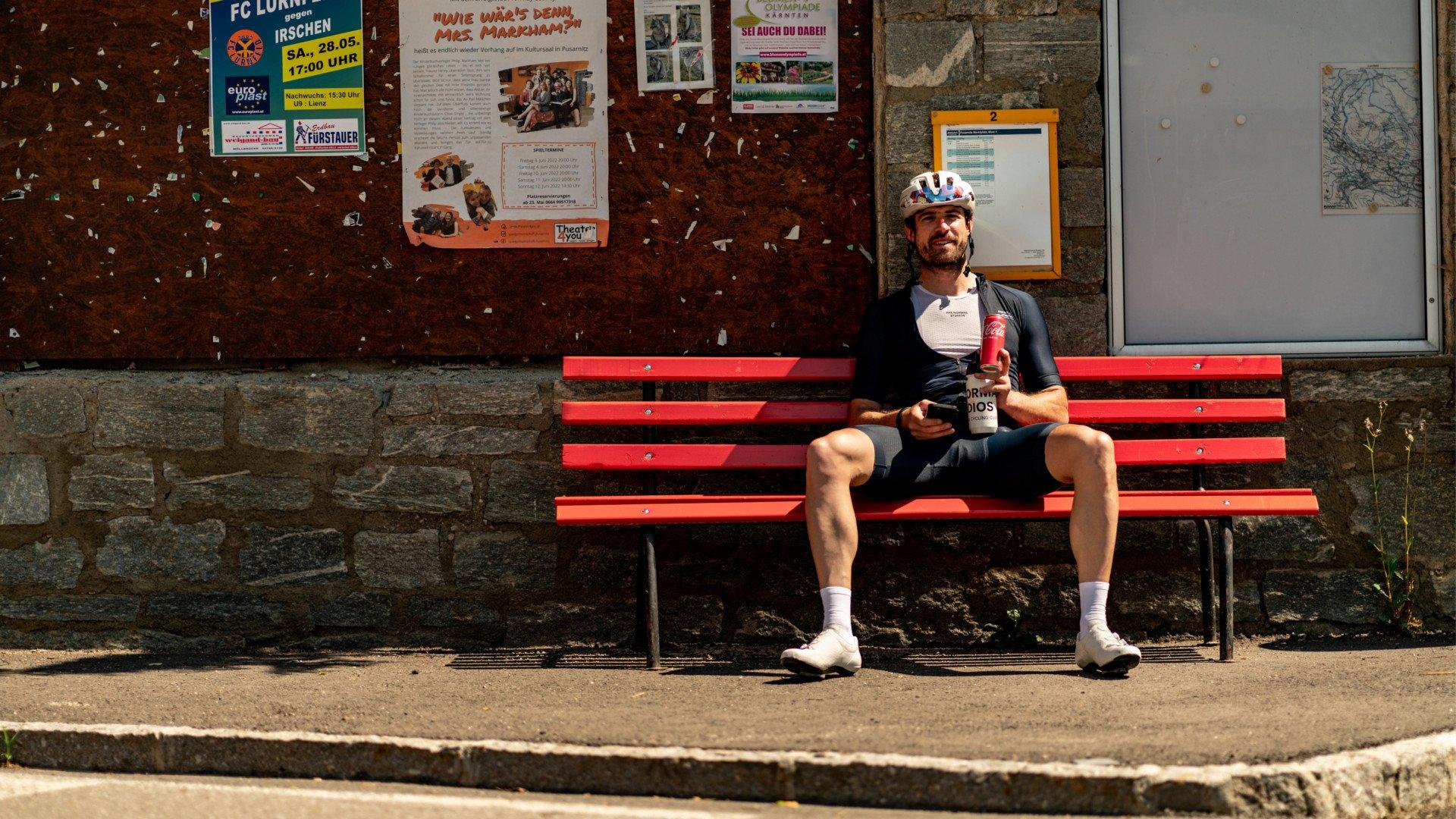 Cyclist wearing white helmet and dark jersey sitting on red bench with beverage after ride