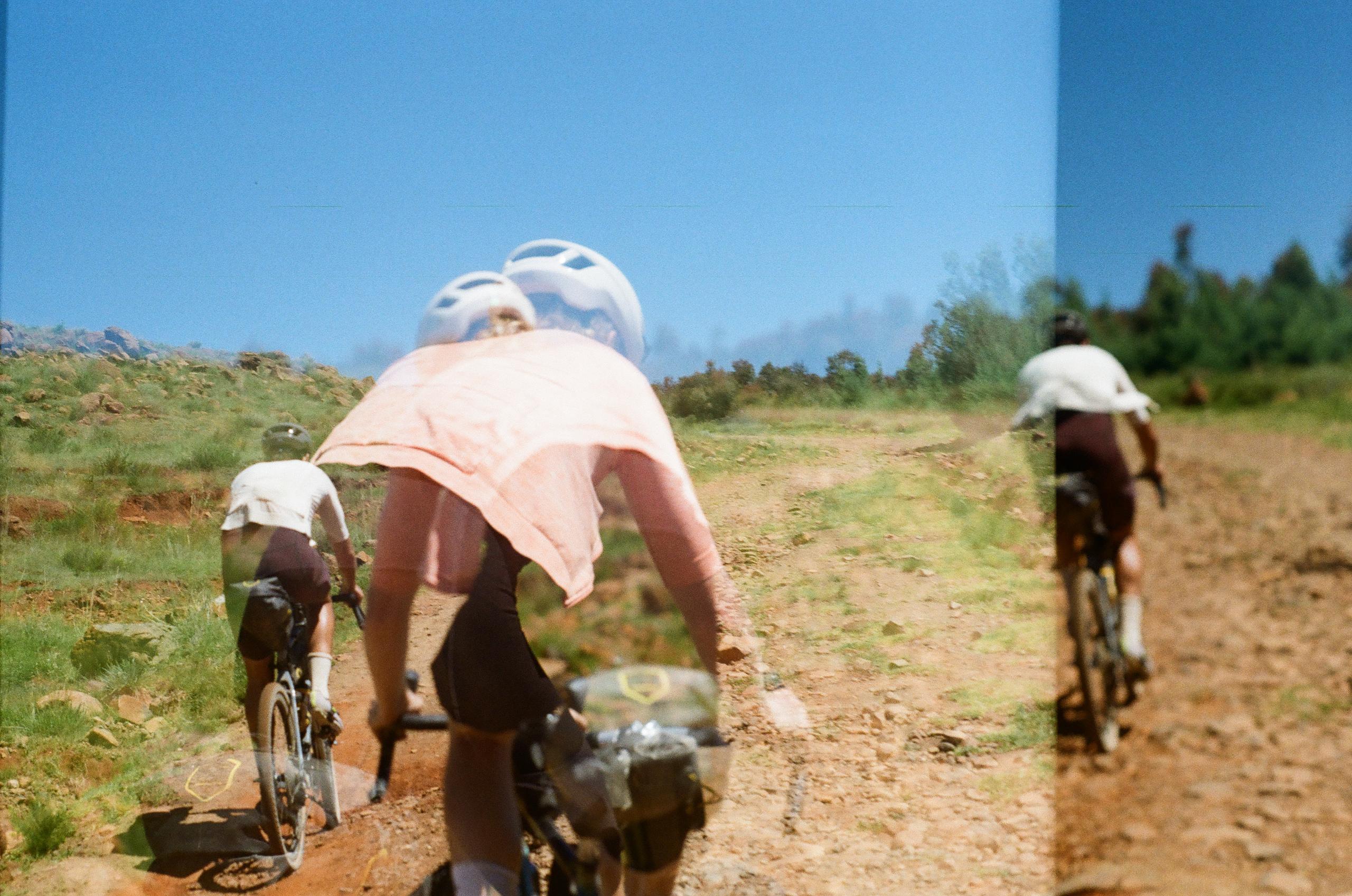 Cyclists in pink jerseys and white caps riding gravel bikes on dirt road through rural landscape