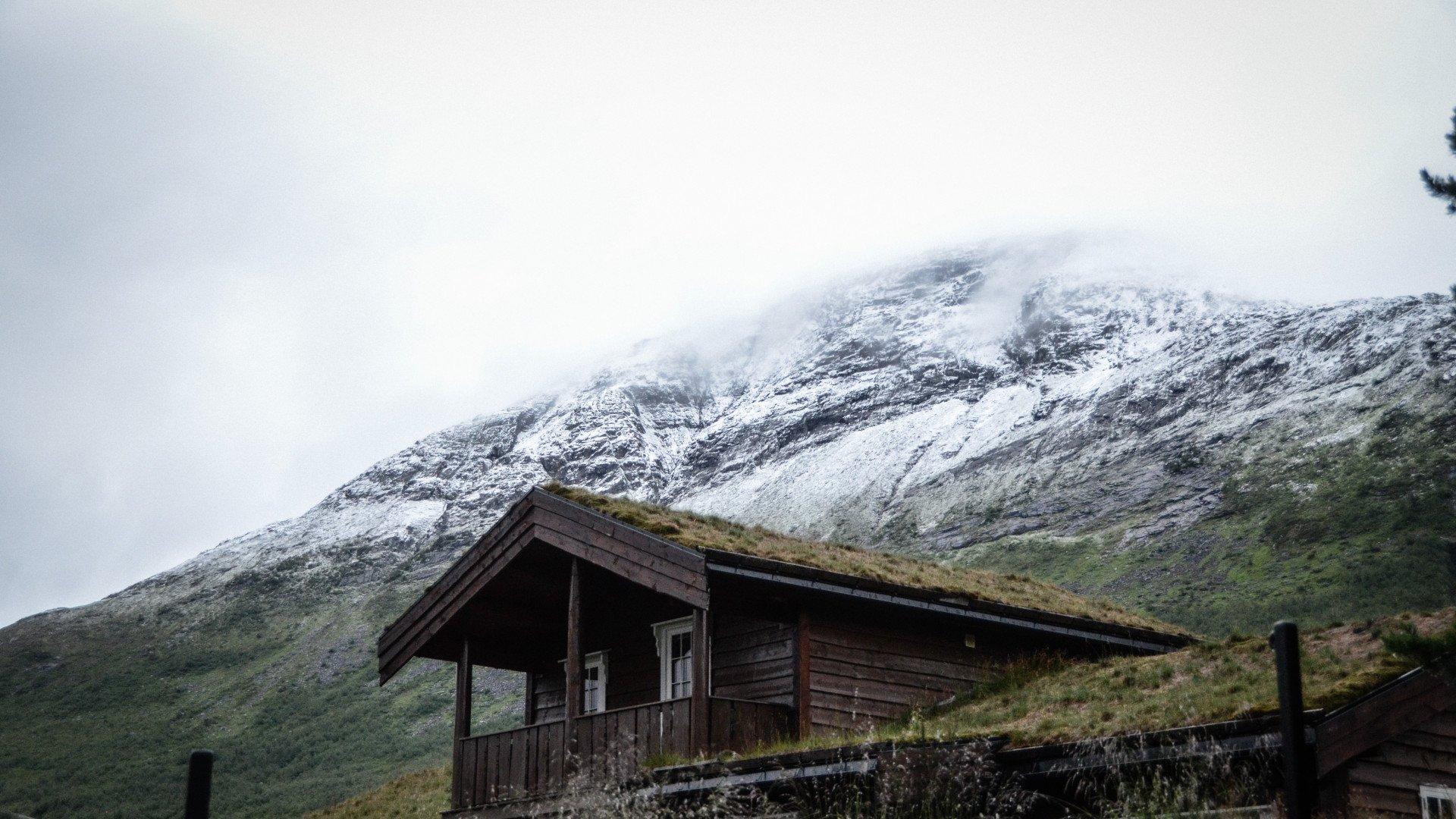 Traditional Norwegian wooden cabin with grass roof against snow-capped mountains in misty weather