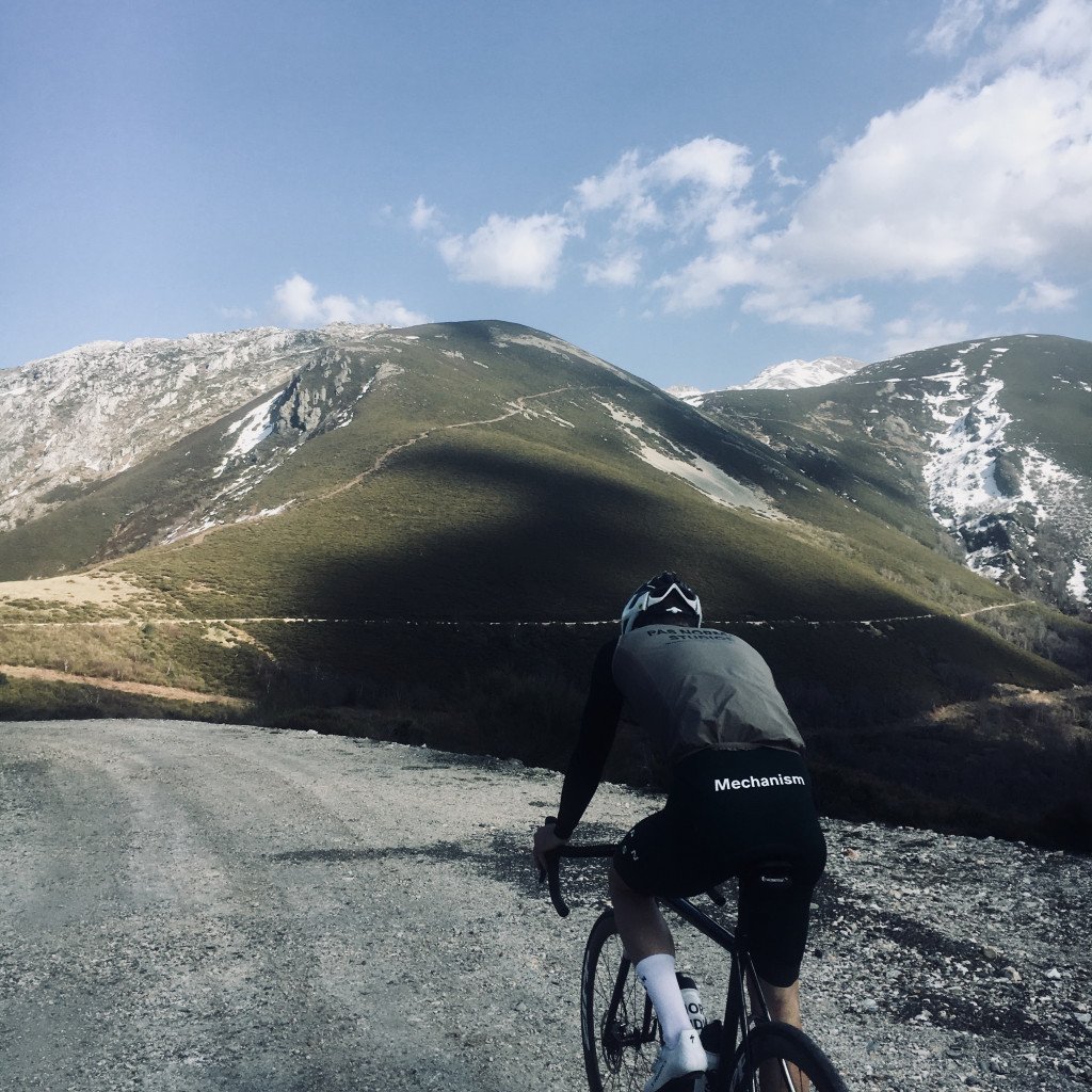 Model wearing dark grey Mechanism jersey cycling through mountain landscape with snow-capped peaks