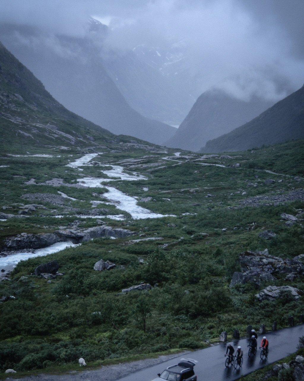 Cyclists on mountain road through Norwegian valley with stream and misty peaks
