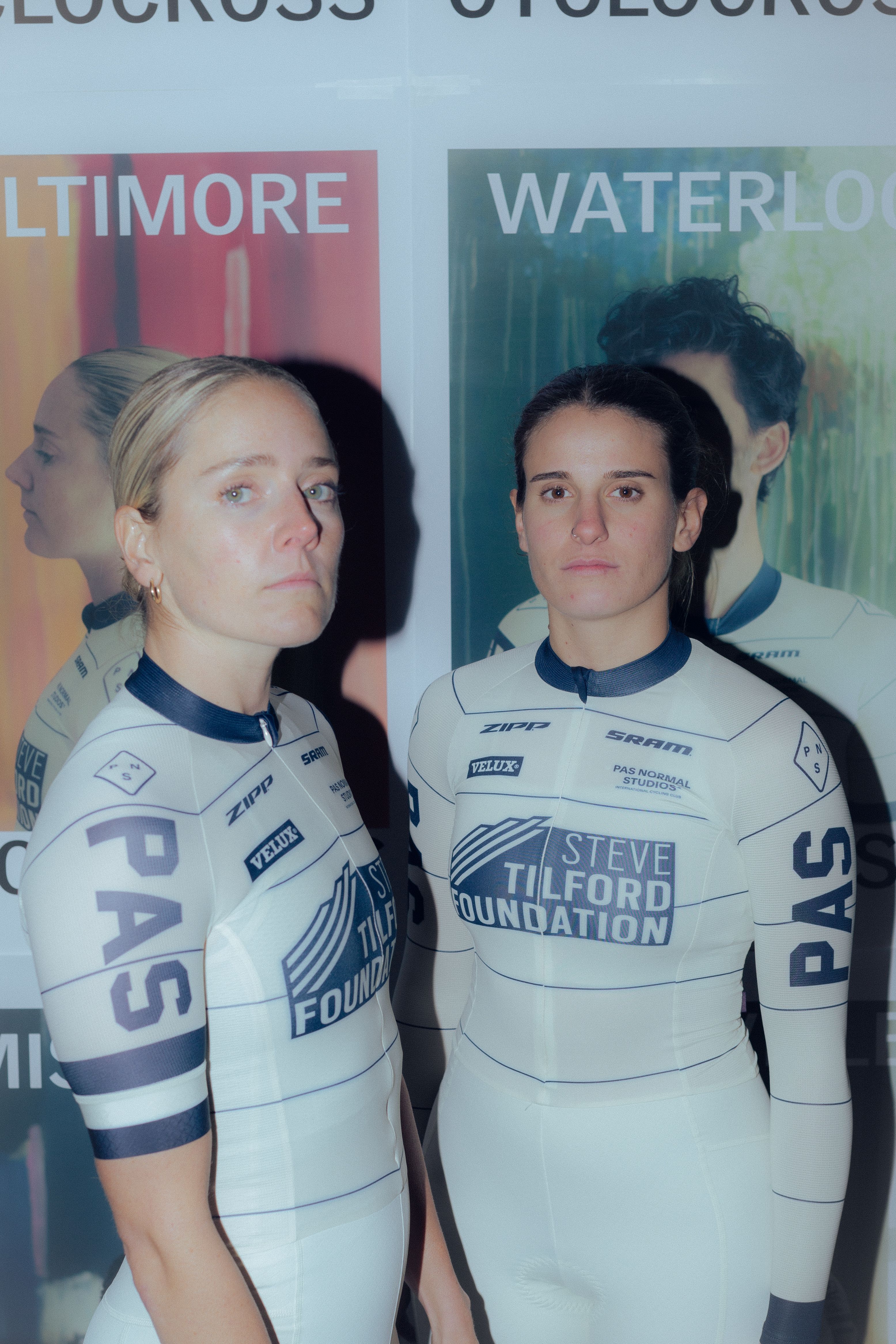 Two female cyclists wearing white team jerseys with blue accents and sponsor logos