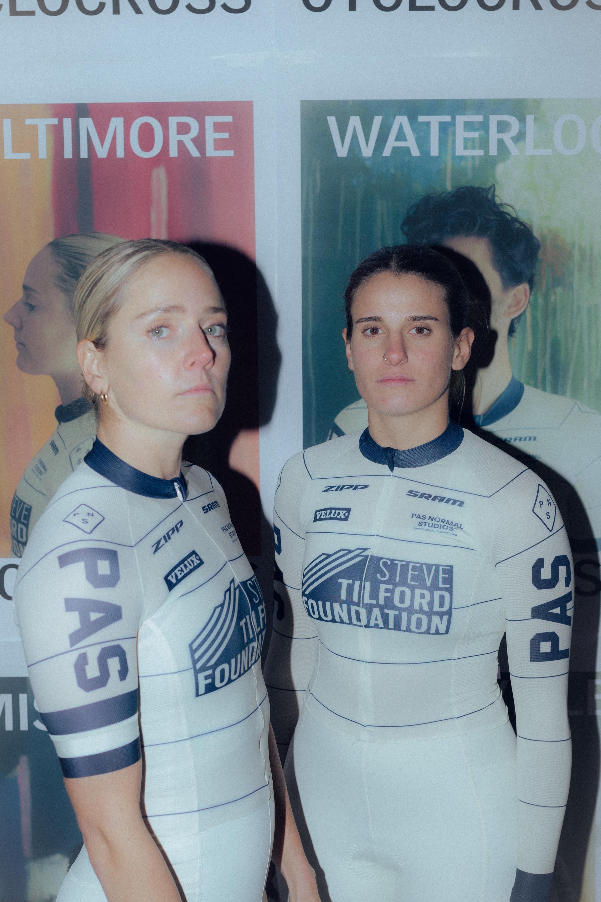 Two female cyclists wearing white team jerseys with blue accents and sponsor logos