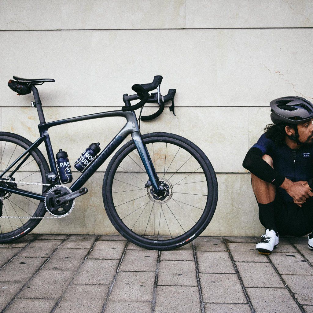 Cyclist in dark cycling kit resting beside road bike against concrete wall