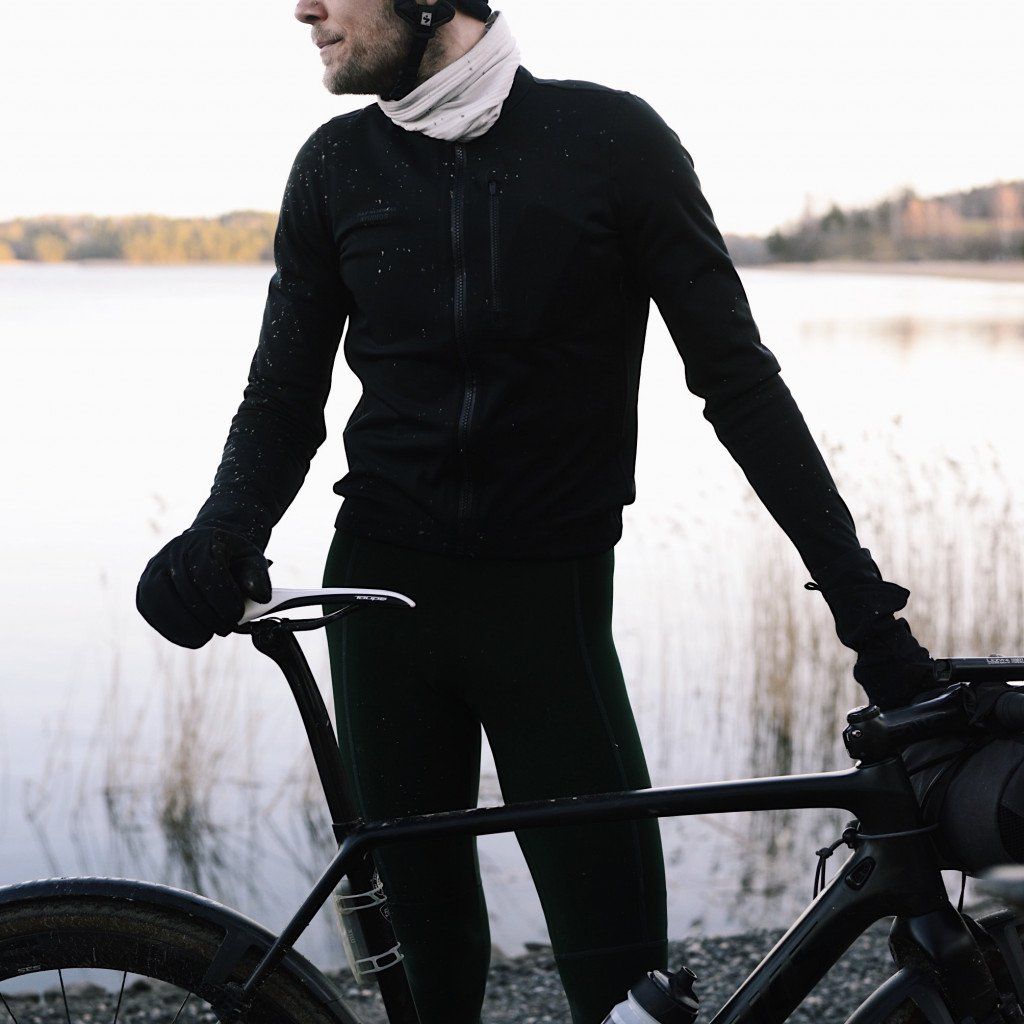 Model wearing black cycling jacket and tights with water droplets, holding bicycle by lake