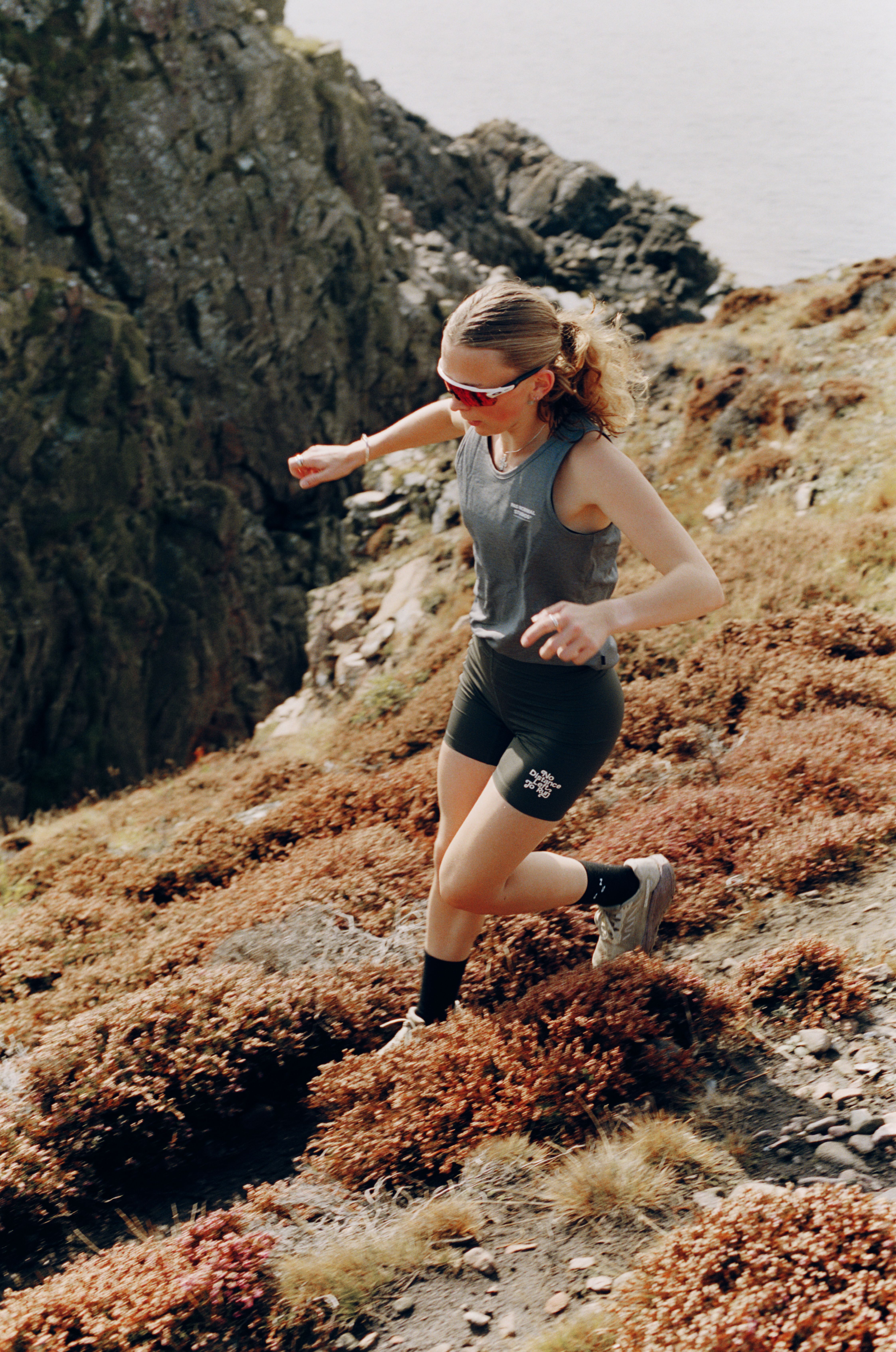 Model wearing grey sleeveless top and black shorts with branding running on rocky terrain