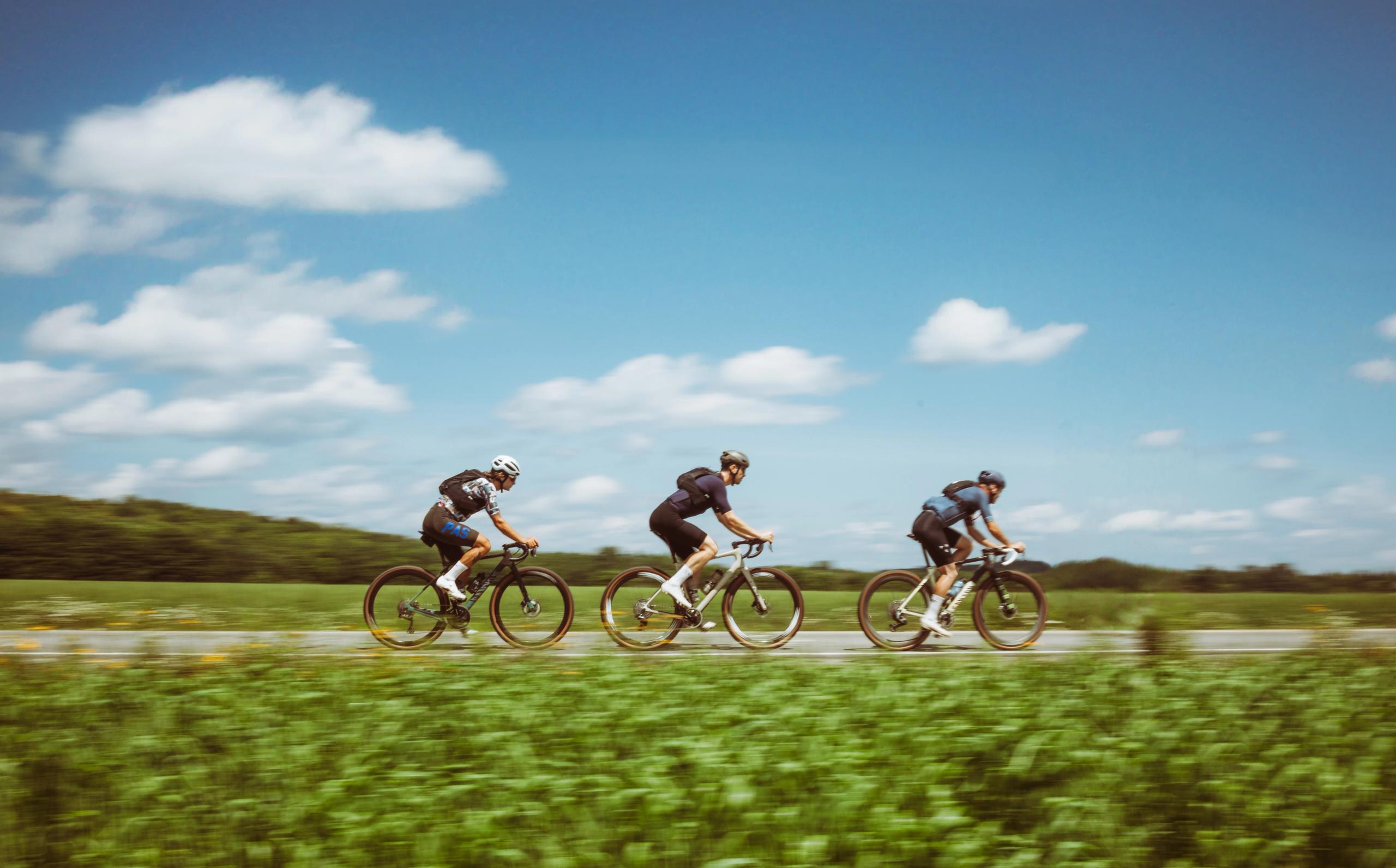 Three cyclists riding time trial bikes on rural road with blue sky and green countryside