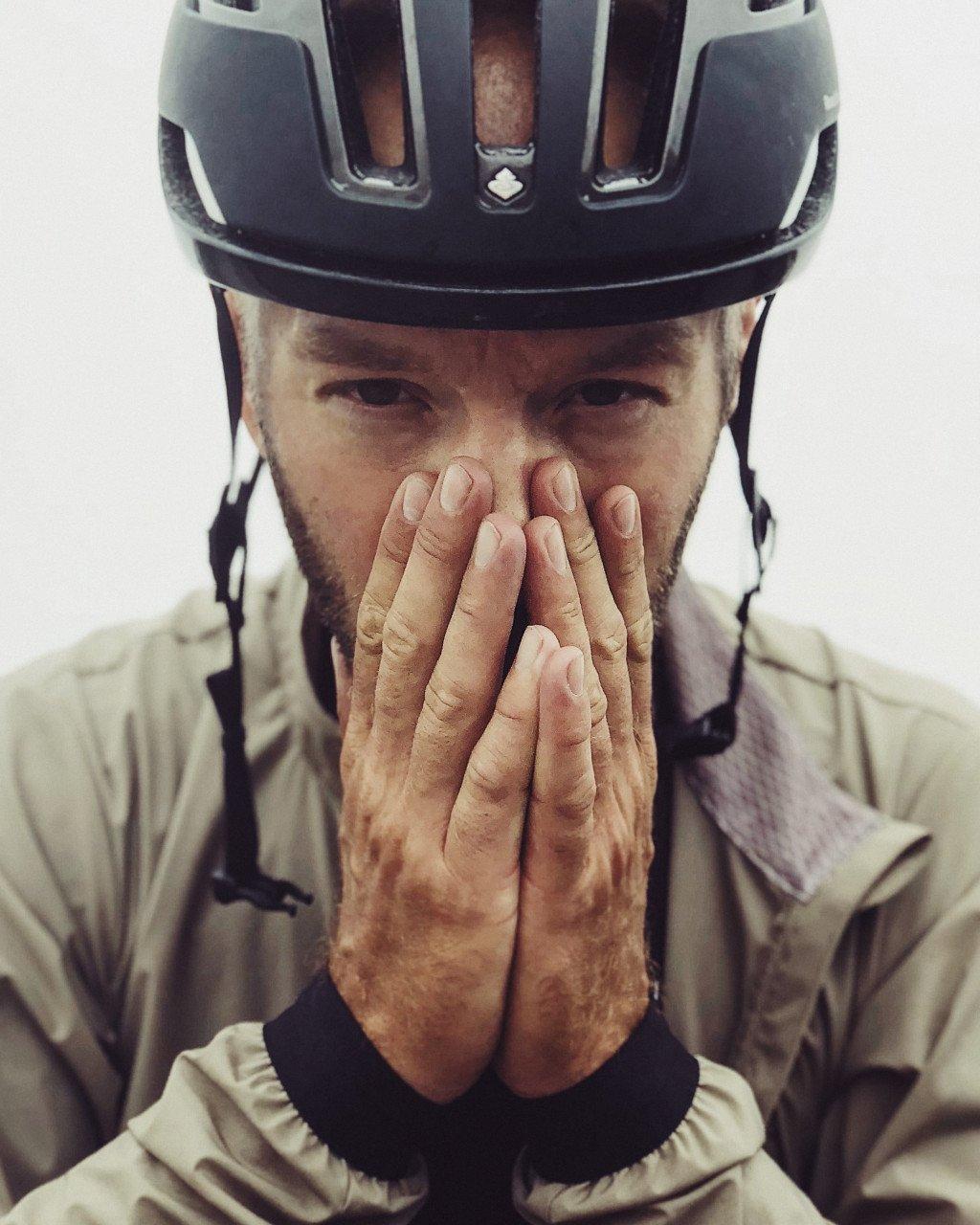 Model wearing cream cycling jacket with black helmet, hands covering face after ride