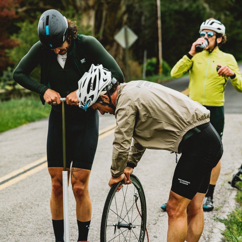 Cyclists on road wearing green skinsuit and beige jacket helping with bike repair during group ride