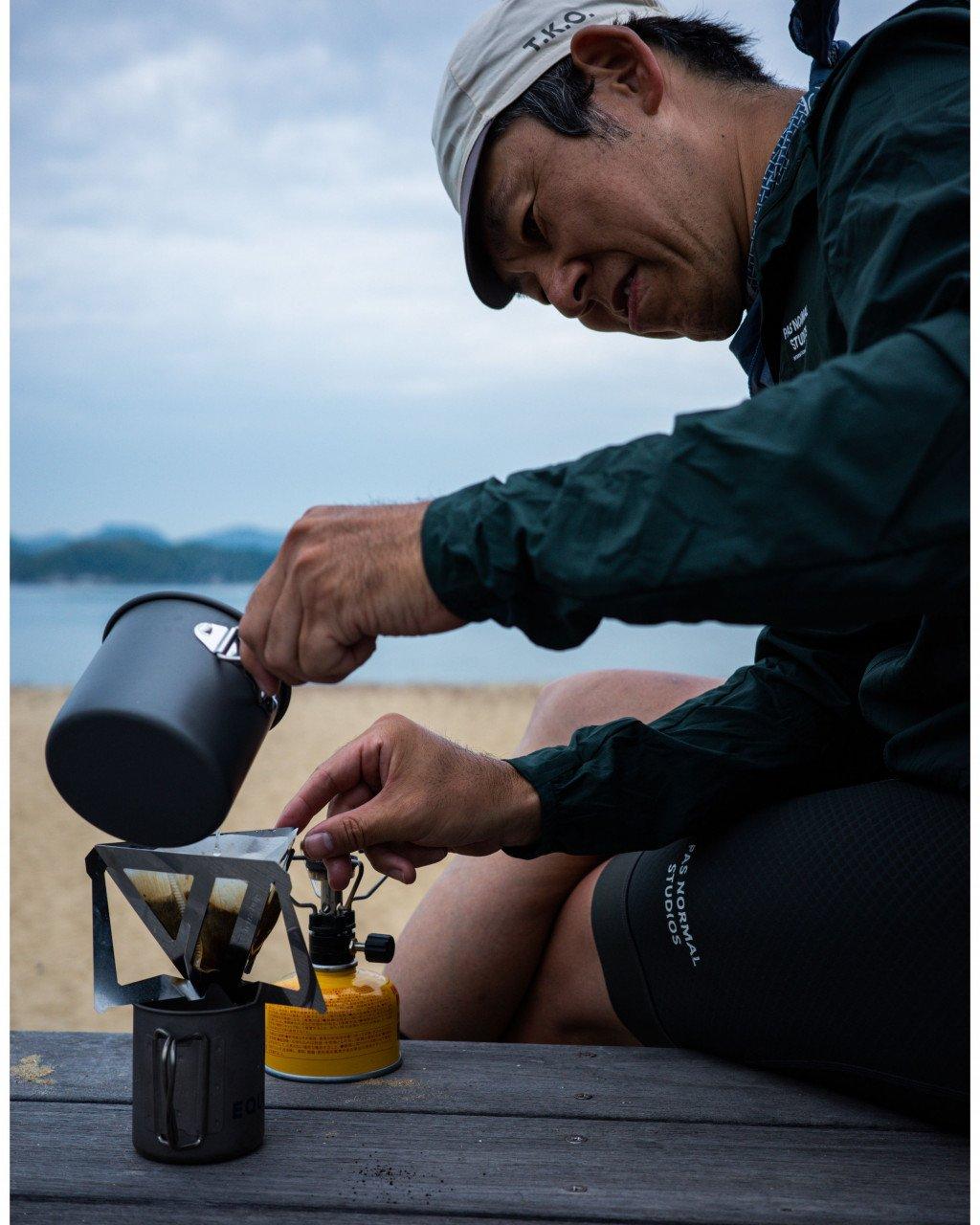 Model wearing green cycling jacket and cap preparing coffee outdoors with camping gear by water