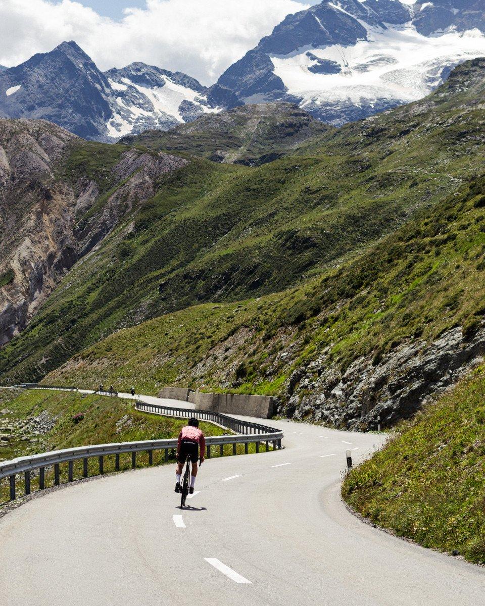 Cyclist in red jersey climbing mountain road with snow-capped Alpine peaks in background
