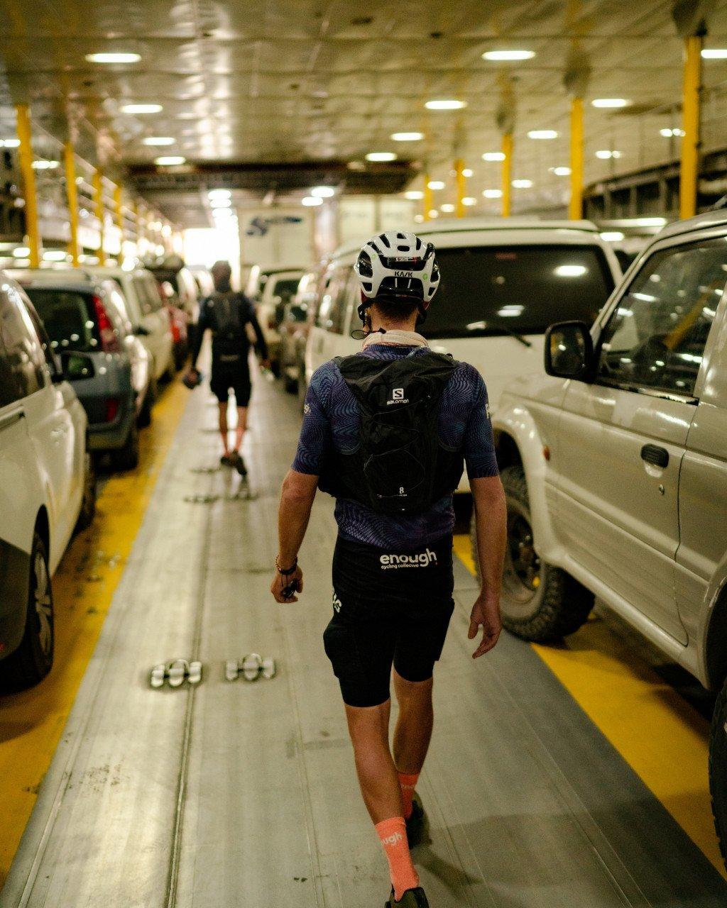 Cyclist wearing navy patterned jersey and black shorts with backpack walking through ferry car deck