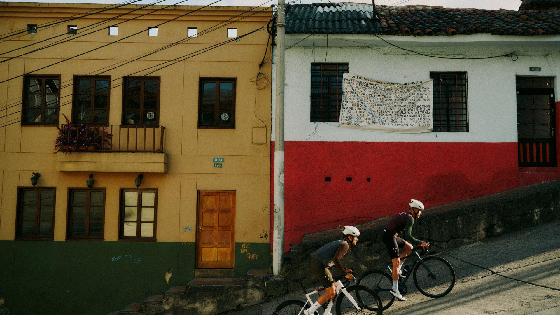 Two cyclists in white helmets riding past colorful colonial buildings on cobblestone street