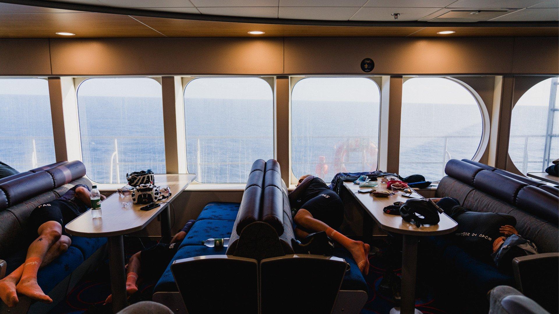 Cyclists resting on ferry benches with cycling gear and ocean view through windows