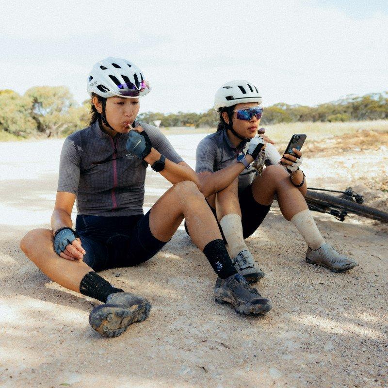 Two cyclists in grey jerseys with purple details taking a break on sandy terrain with bikes nearby