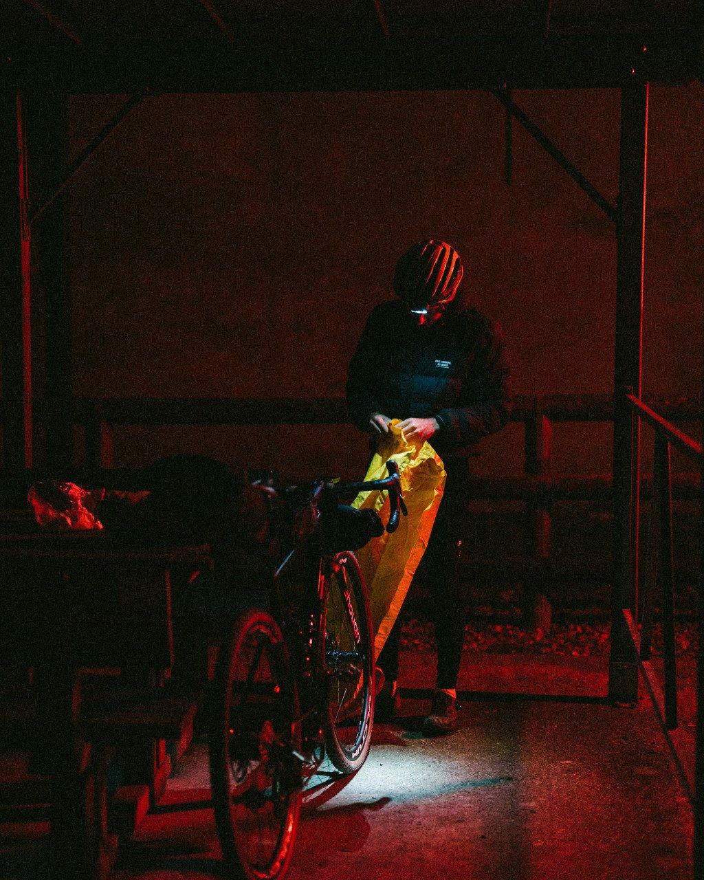 Model wearing dark cycling jacket and yellow pants with helmet, standing with bike in dimly lit industrial space