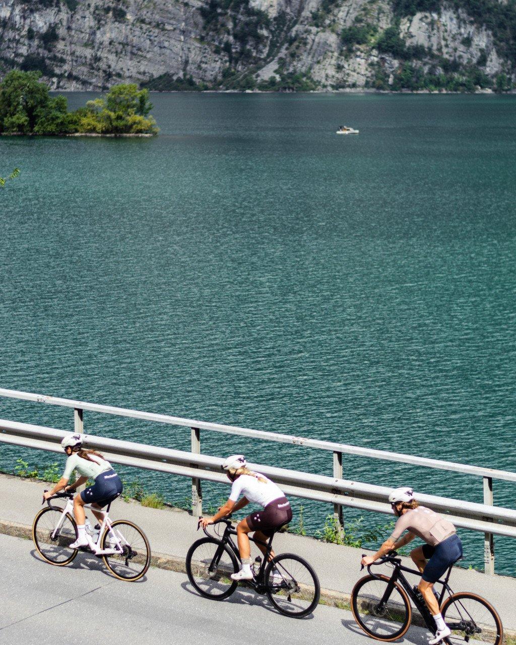 Three cyclists in aerodynamic position riding along lakeside road with mountains in background