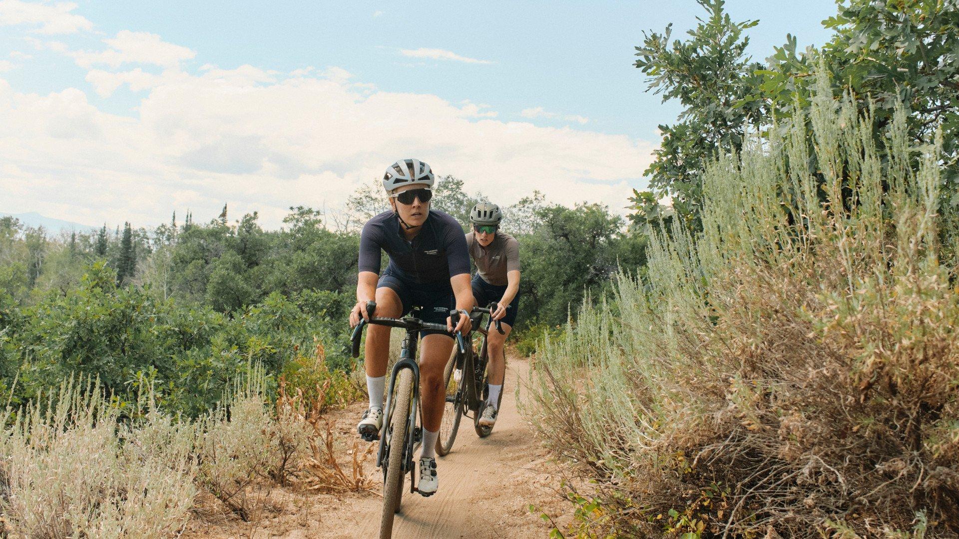 Two cyclists riding gravel bikes on dirt trail through natural landscape with trees and vegetation