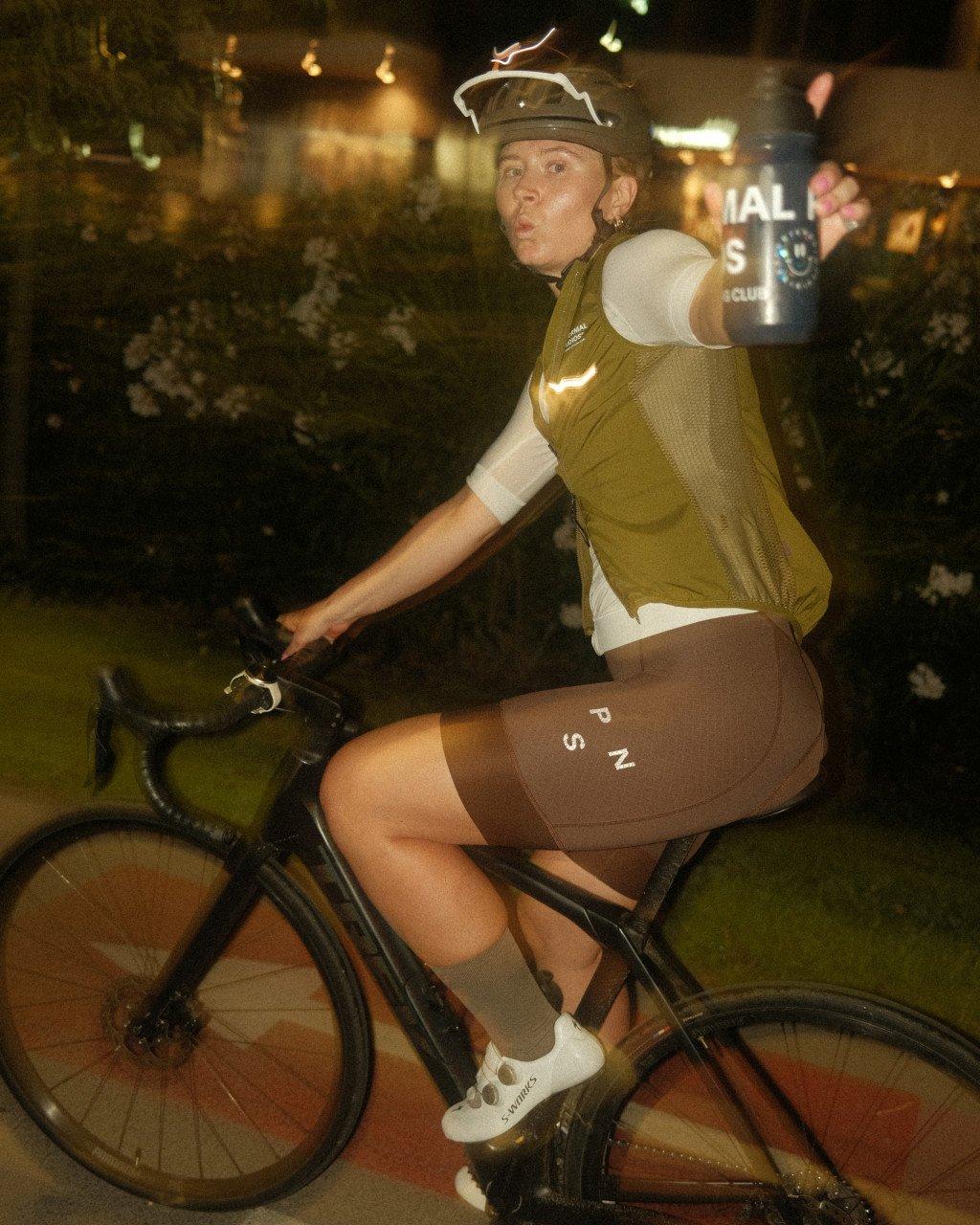 Model wearing olive green cycling vest and brown shorts with branding on indoor bike trainer