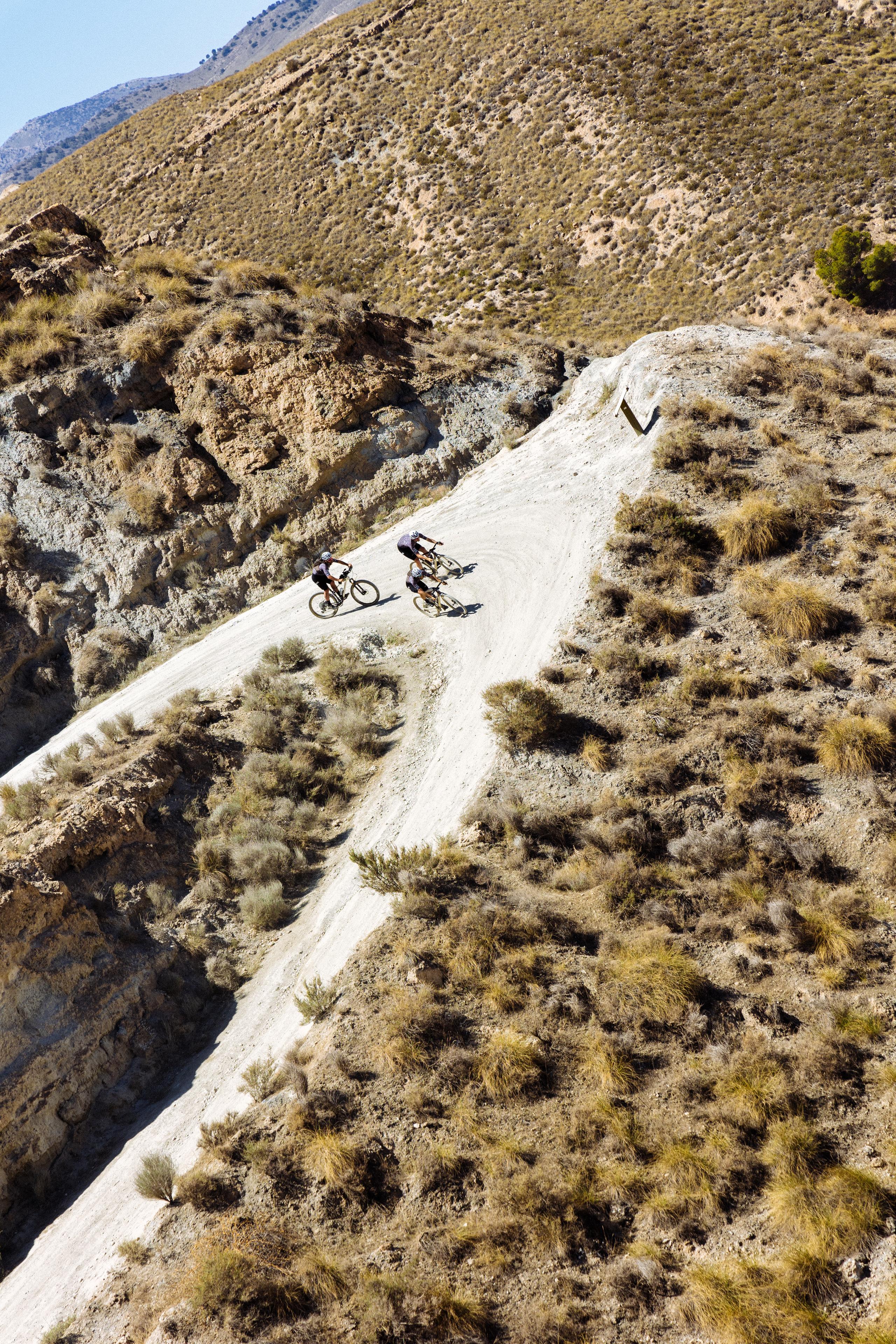 Two cyclists riding on mountain road through arid landscape with rocky terrain and sparse vegetation