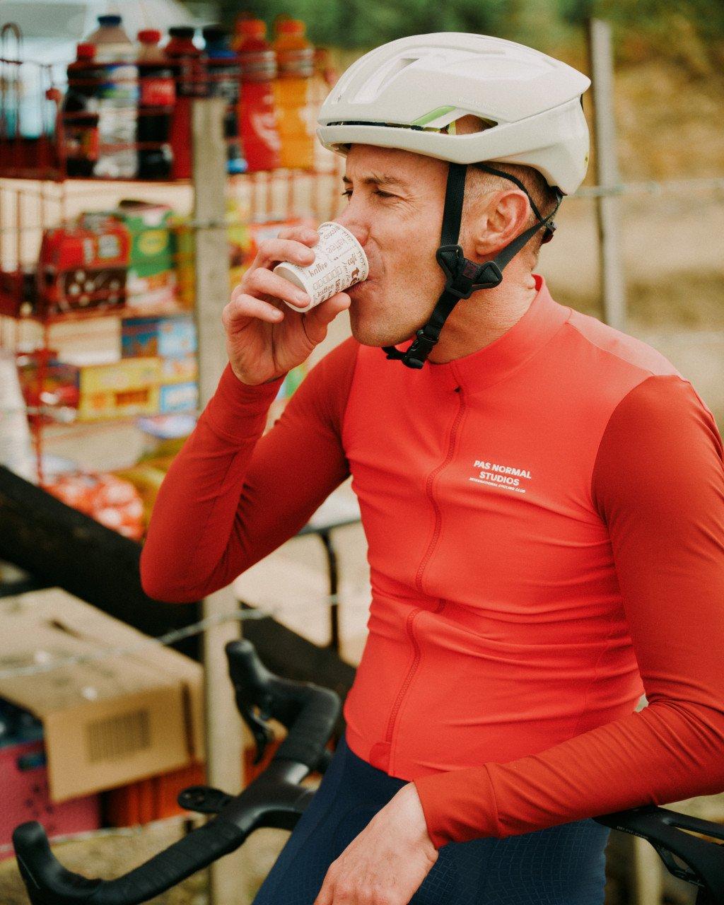 Model wearing red long sleeve cycling jersey with chest logo drinking at cafe stop