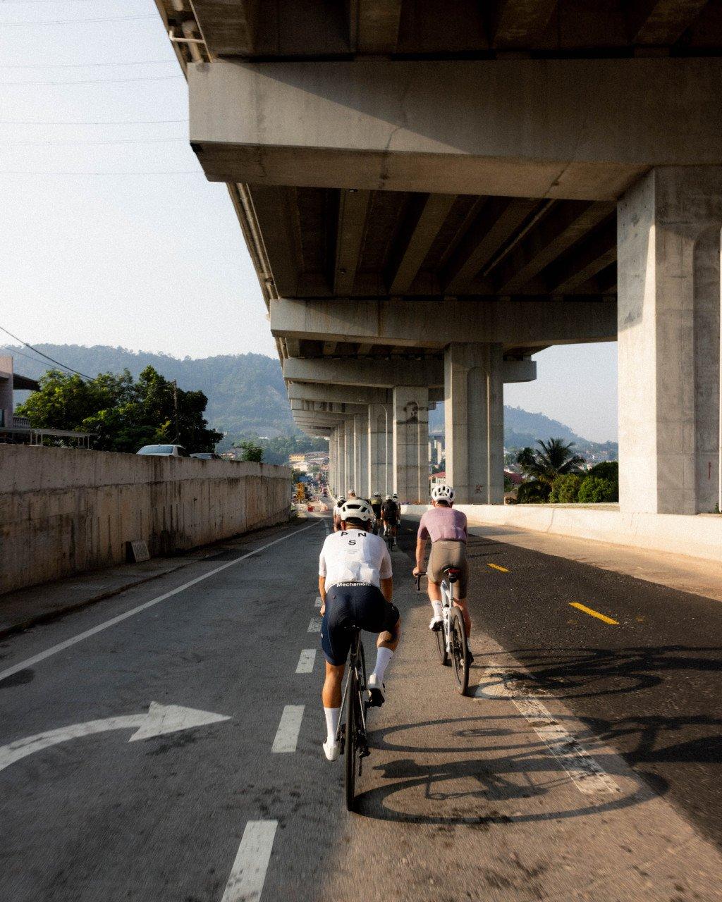 Cyclists riding under concrete bridge overpass in tropical setting with mountains in background