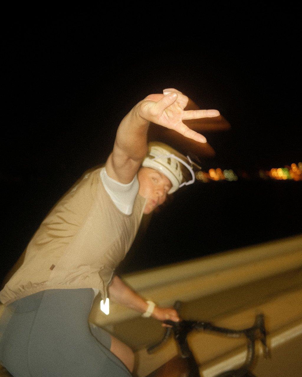 Model wearing cream long-sleeve jersey making hand gesture while cycling at night