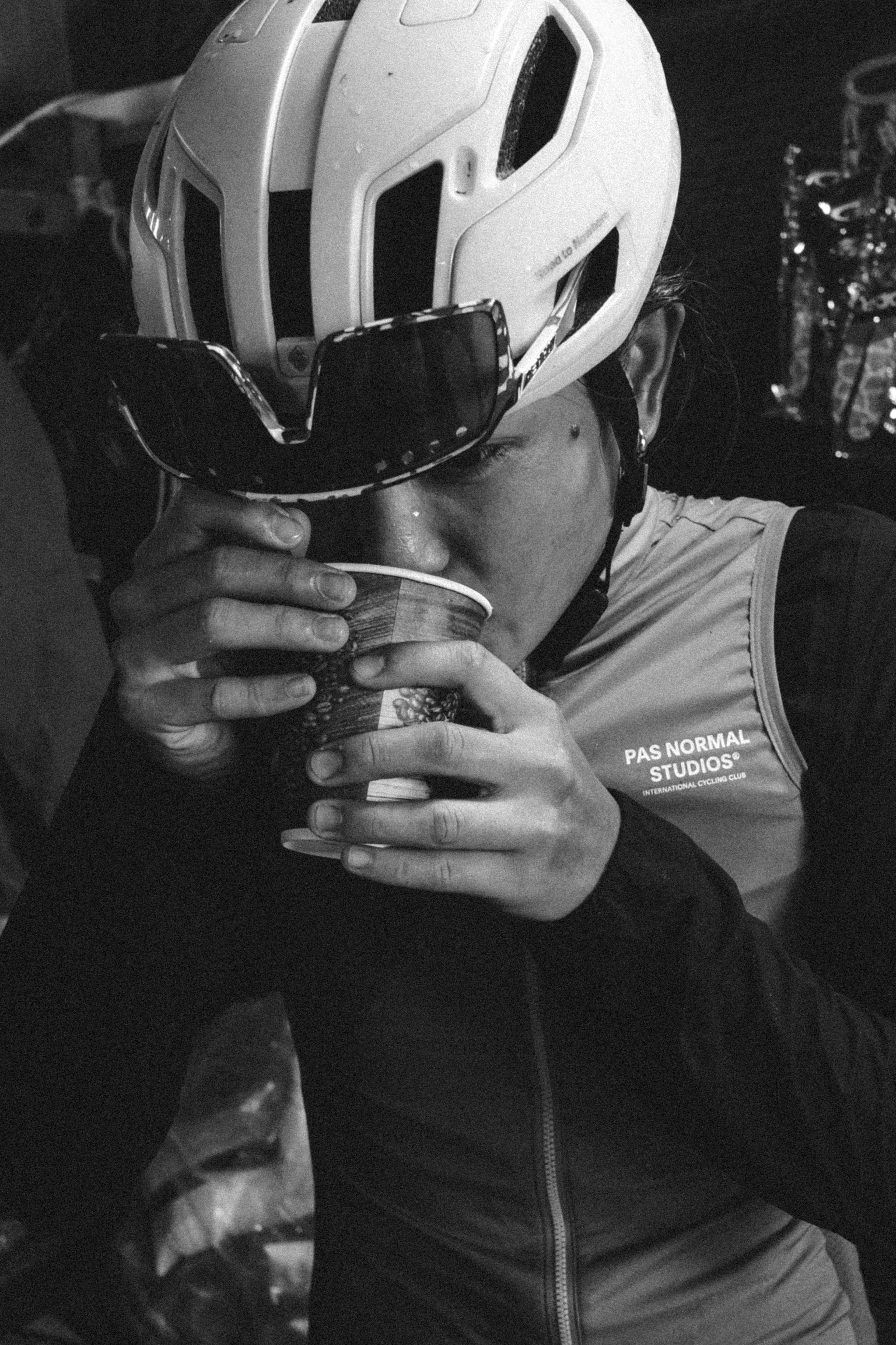 Cyclist in white helmet and sunglasses drinking from glass, wearing cycling apparel with logo