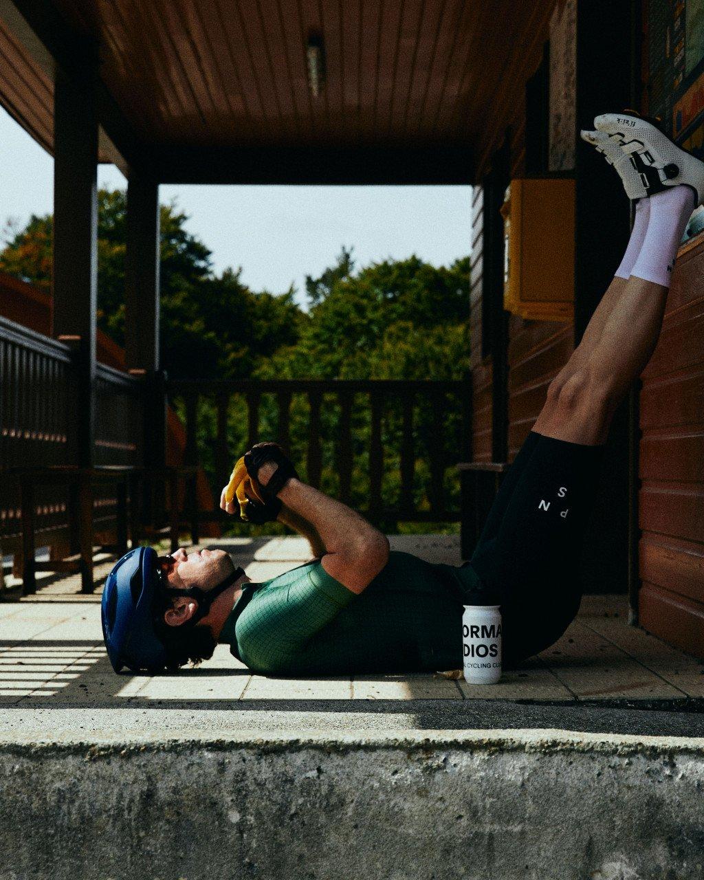 Model wearing green cycling jersey and black bib shorts lying on wooden deck with white socks and cycling shoes