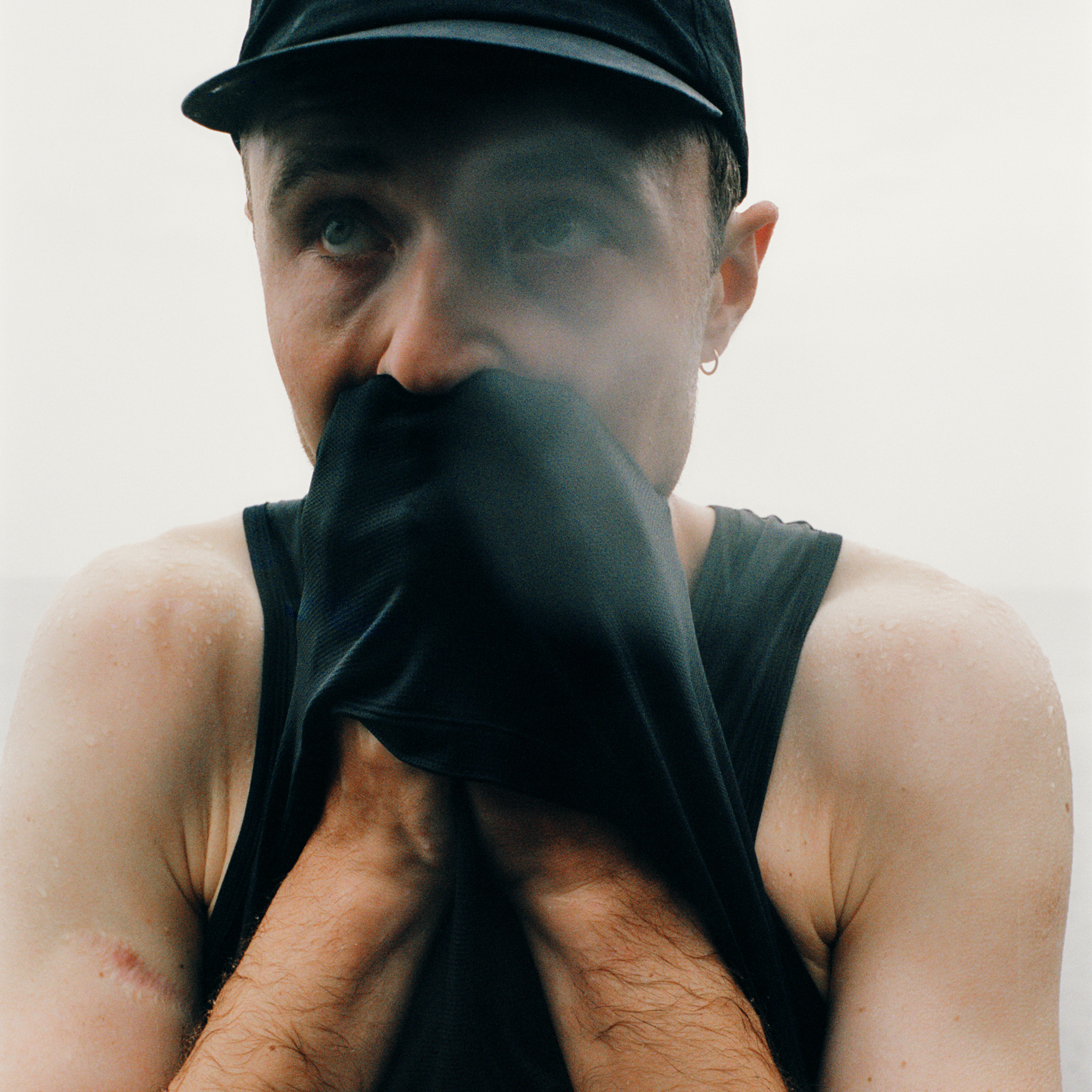Model wearing black cap and face covering with dark tank top, arms raised