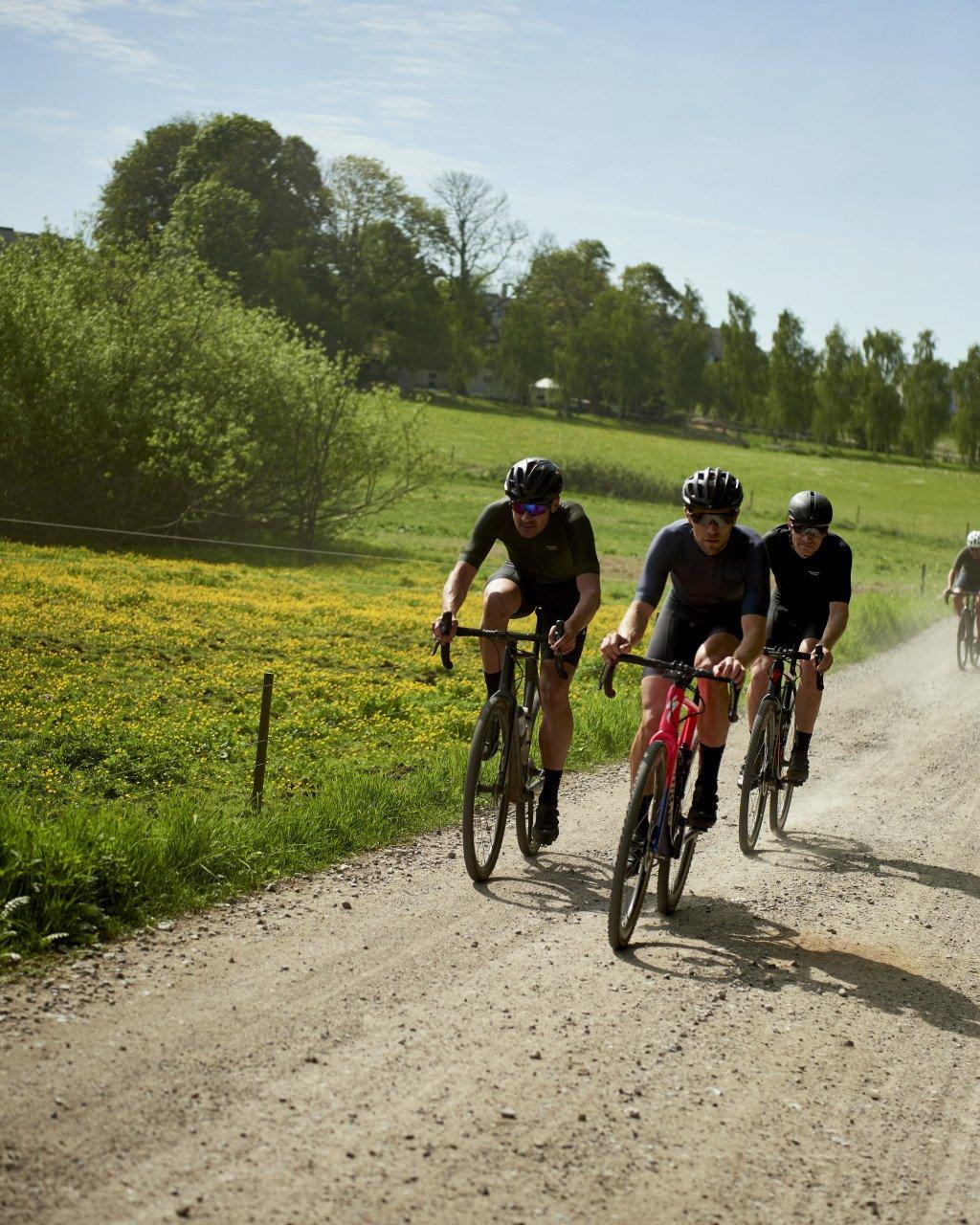 Three cyclists in dark cycling kit riding gravel bikes on countryside path through green fields