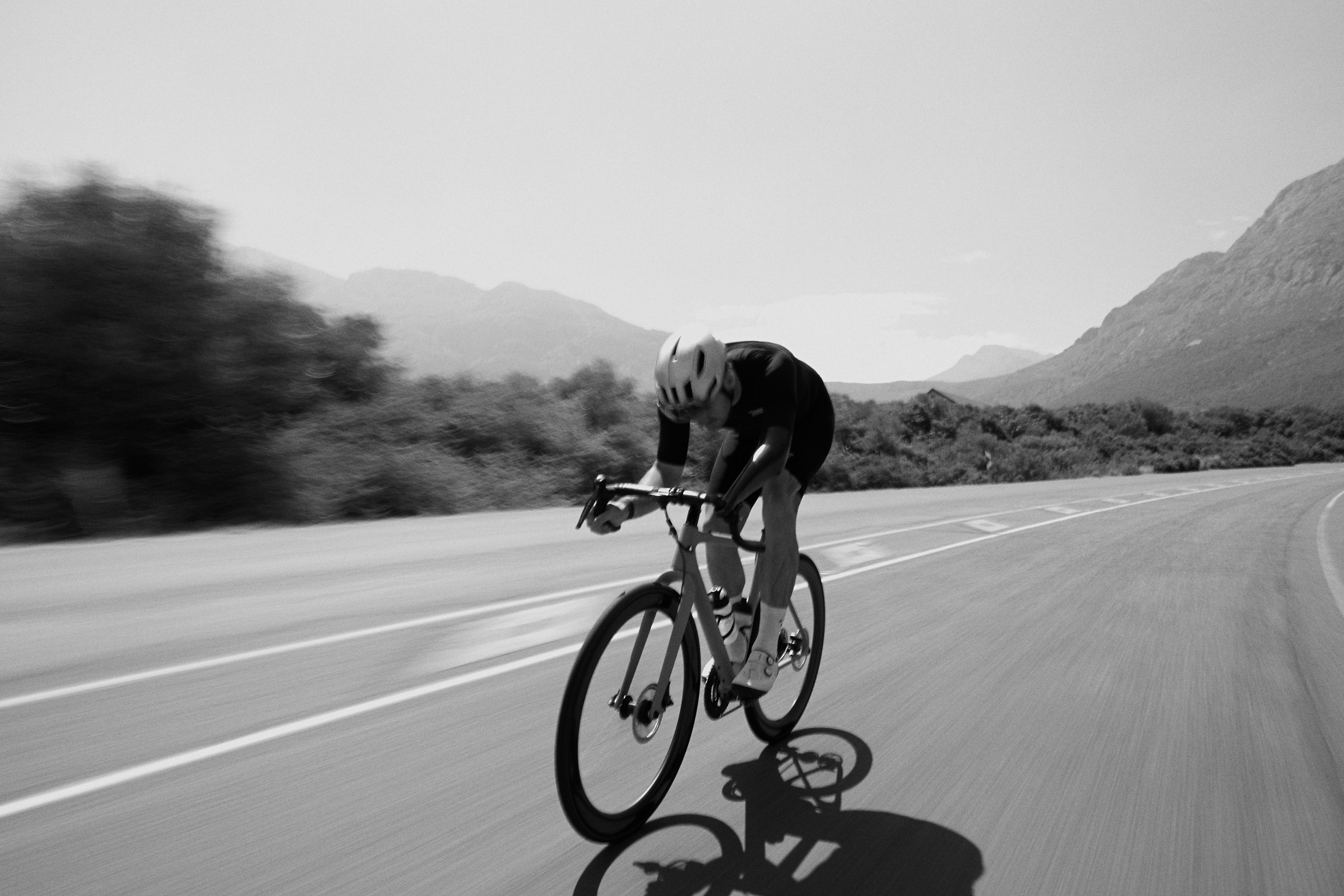 Cyclist in aerodynamic position on time trial bike riding through mountainous landscape in black and white