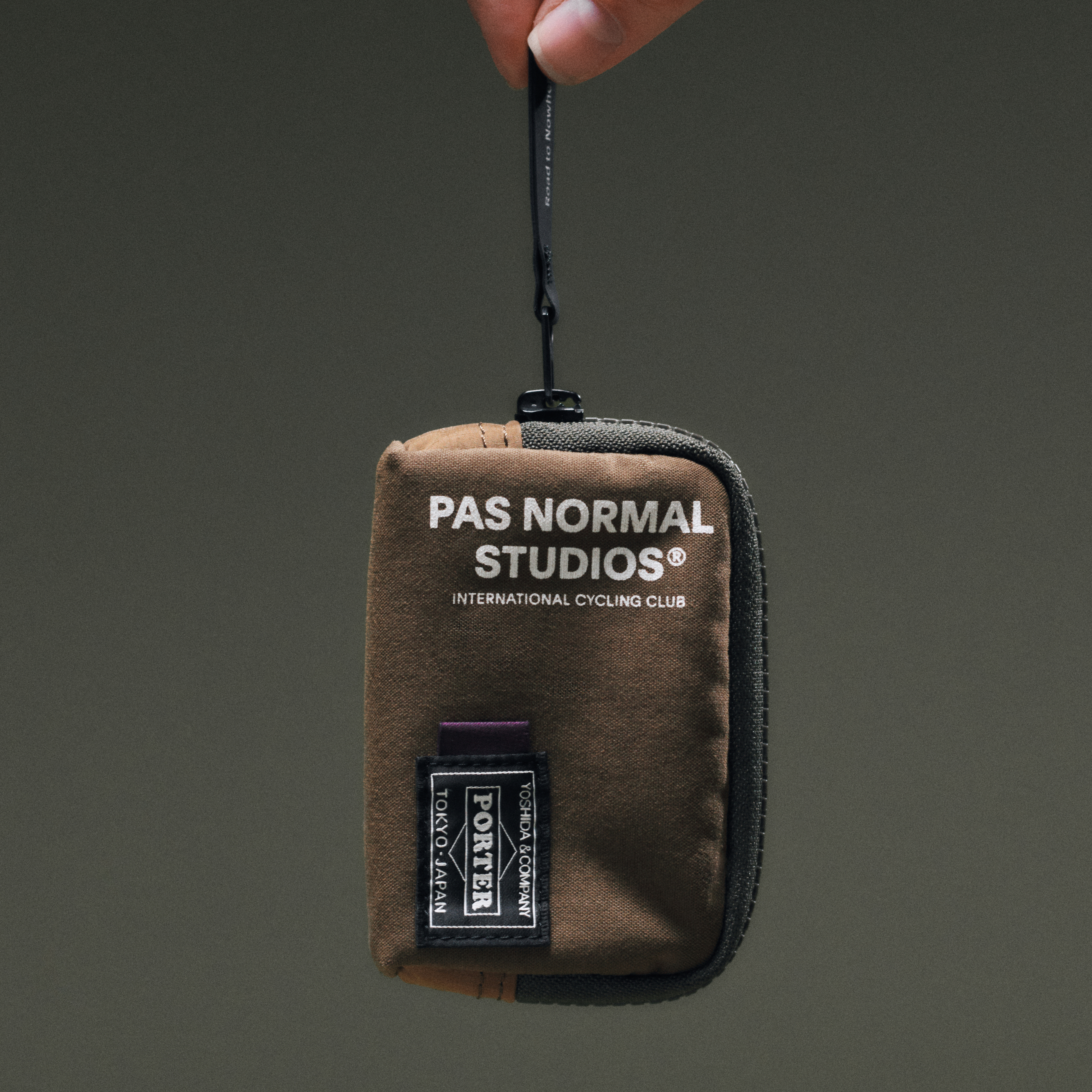Tan Porter-Yoshida coin case held up, front view with branded woven label and black clip hook
