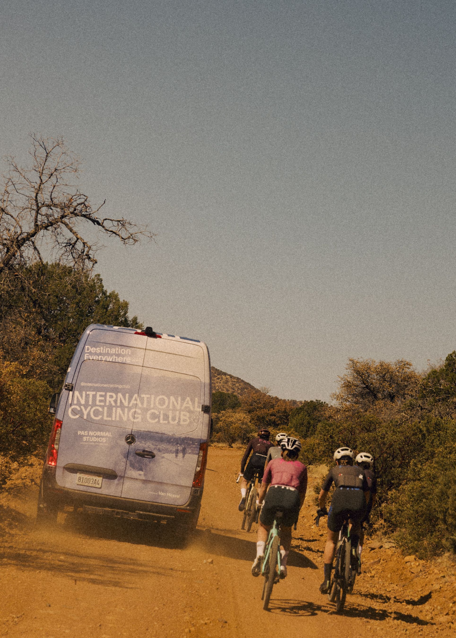 Cyclists riding gravel road alongside support van in desert landscape with hills and sparse vegetation
