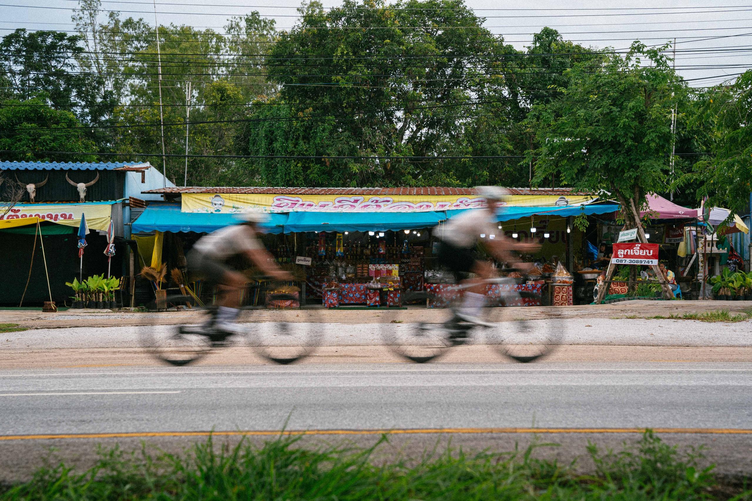 Two cyclists in motion blur riding past colorful Thai roadside market stalls