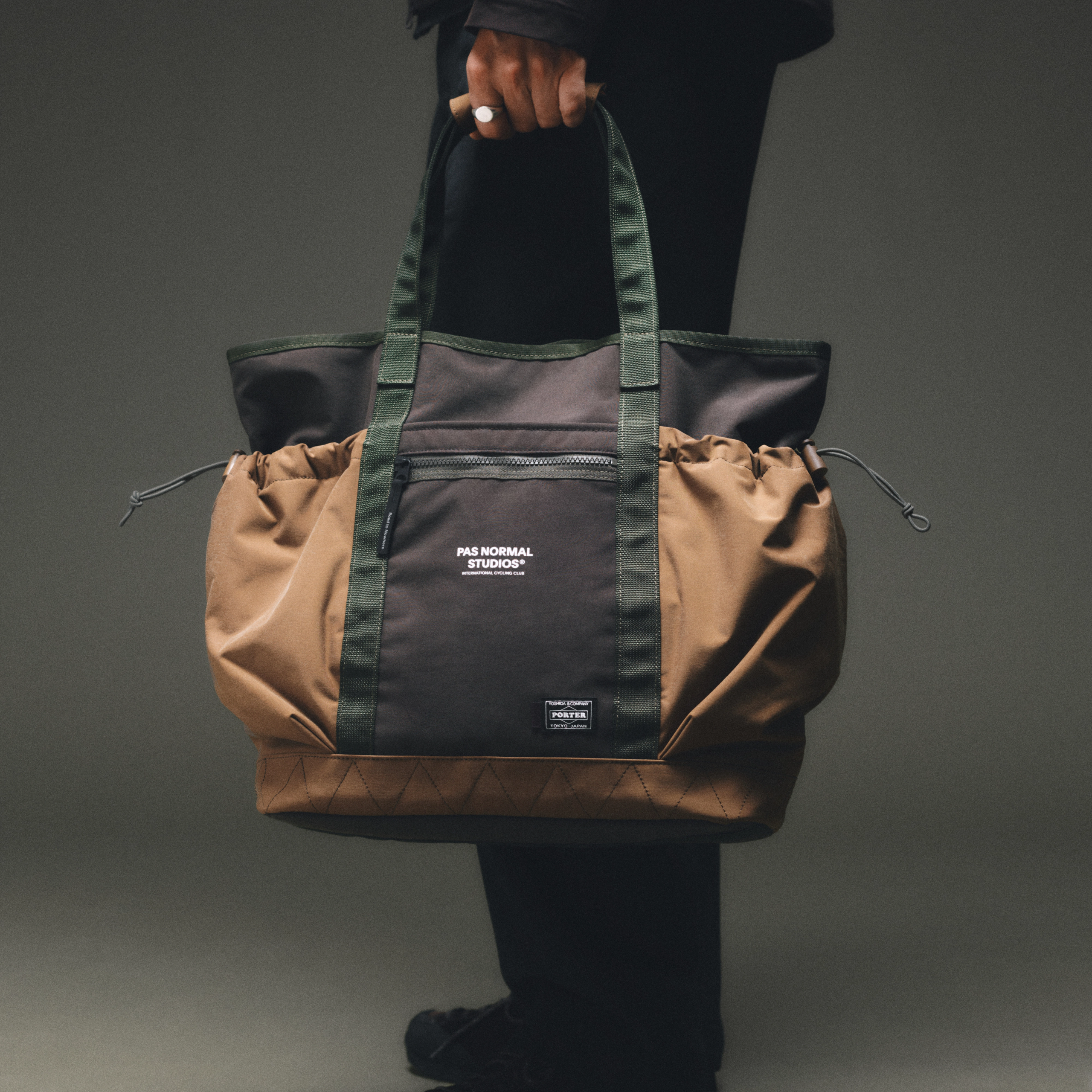 Tan and black Porter-Yoshida tote bag held by model, front view with green webbing straps and drawstring side panels