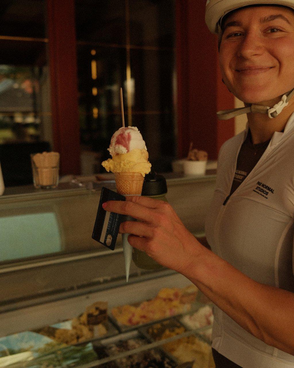 Model wearing white cap and white jersey with chest branding, holding ice cream at gelato shop