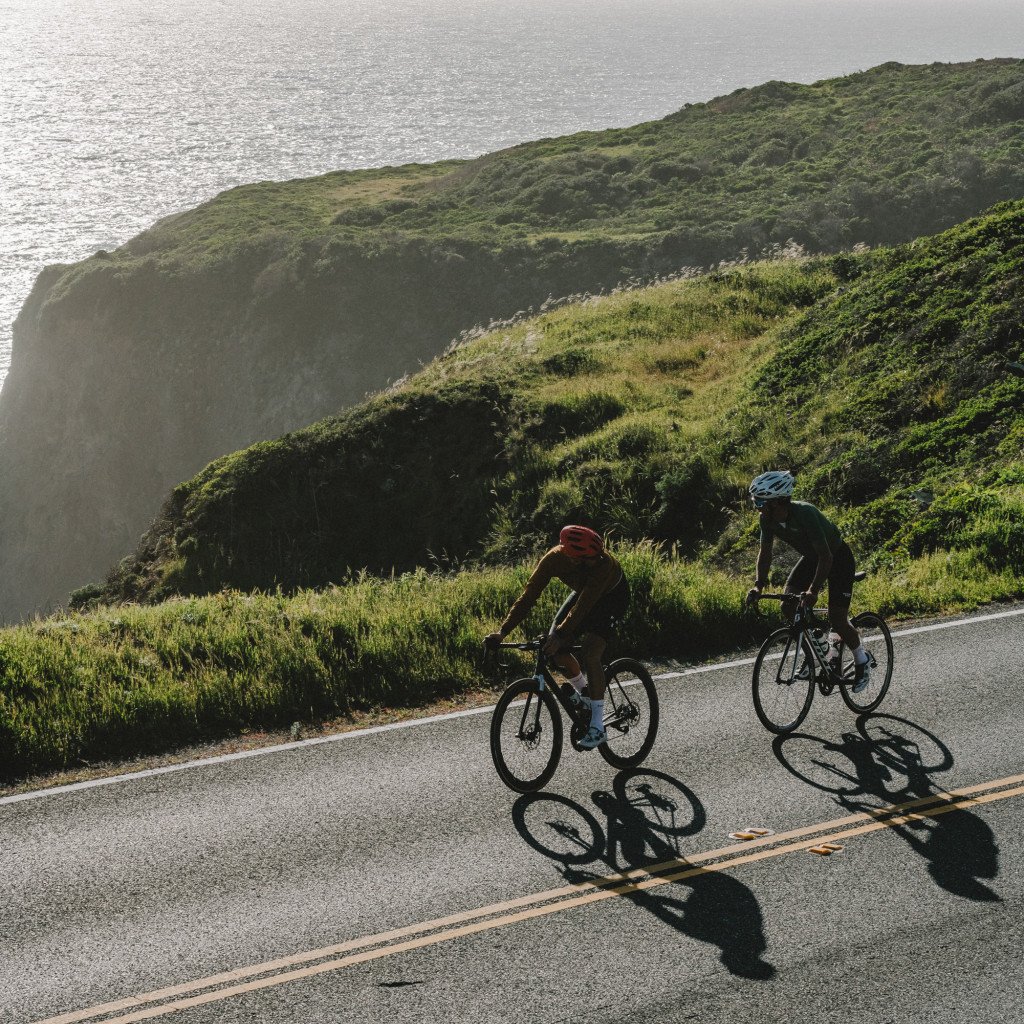 Two cyclists riding coastal road with green hills and ocean backdrop in Sonoma, CA