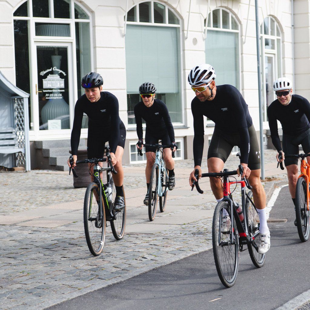 Four cyclists wearing black long sleeve jerseys and helmets riding time trial bikes on cobblestone street