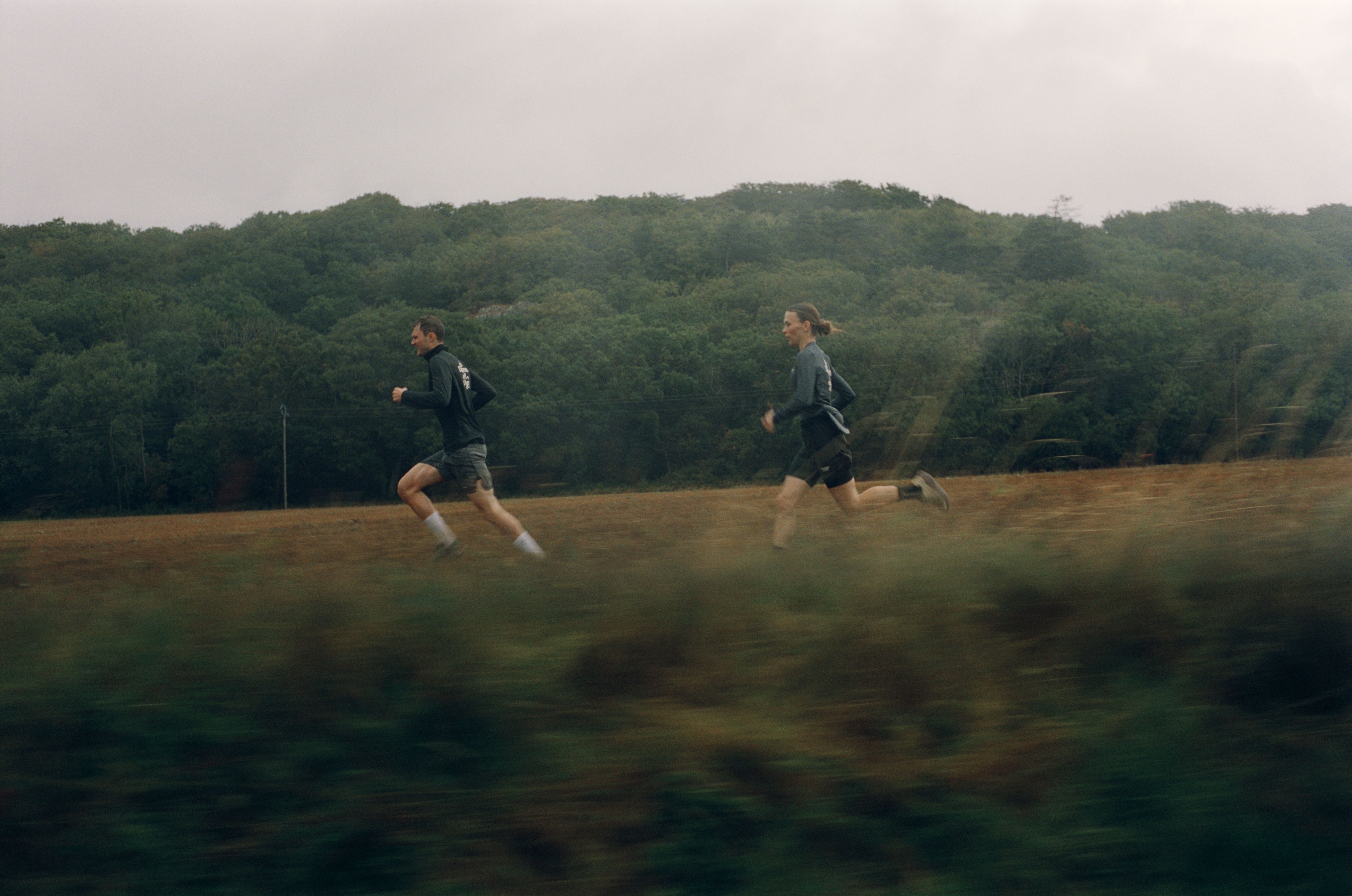 Two runners in dark athletic wear running across open field with forested hills in background