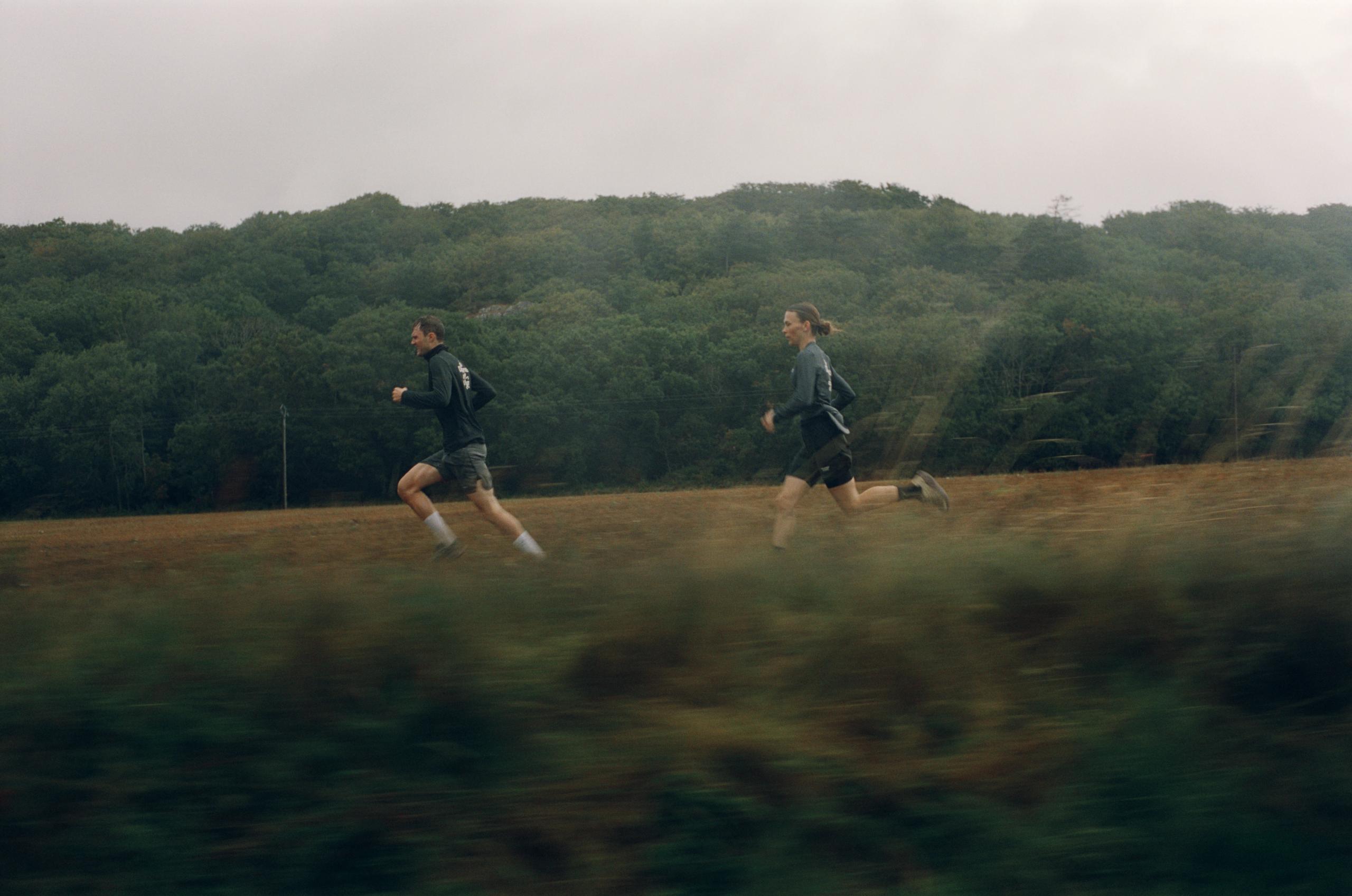 Two runners in dark athletic wear running across open field with forested hills in background