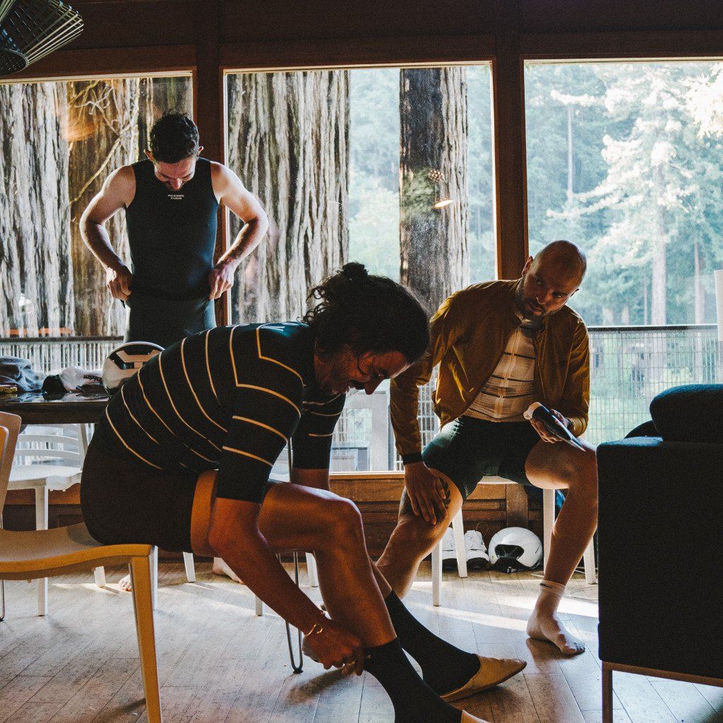 Three cyclists preparing in cabin interior, one wearing striped jersey putting on shoes, others in casual wear