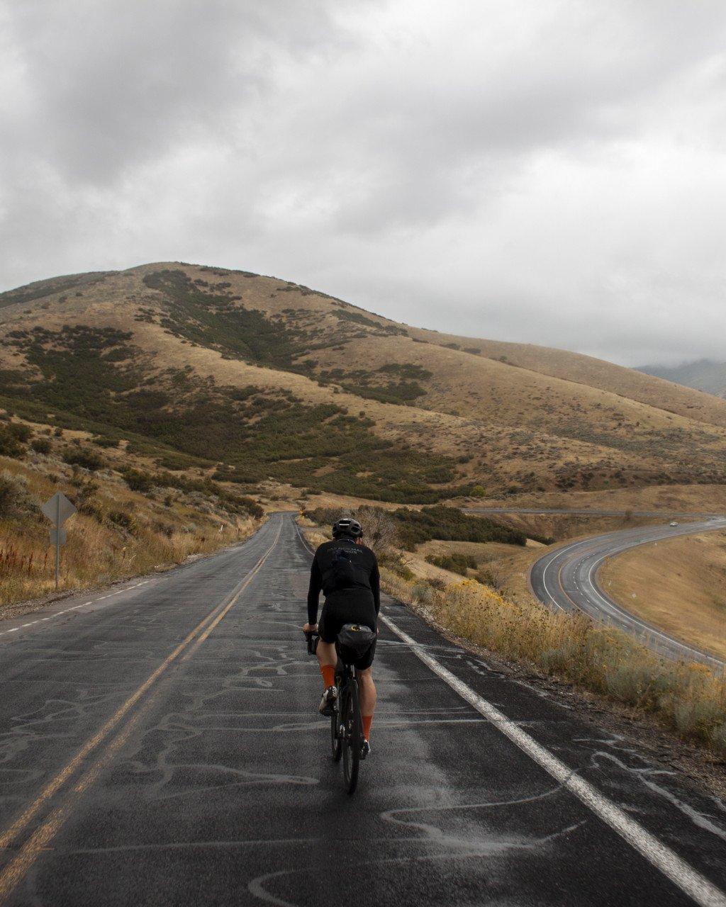 Cyclist riding away on empty road through rolling hills under overcast sky