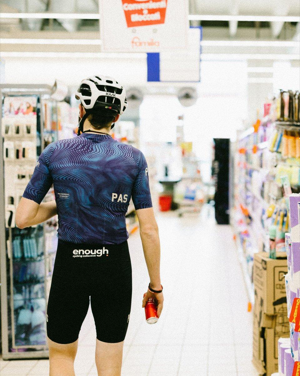 Model wearing navy PAS jersey with wave pattern and black shorts, standing in convenience store aisle with white helmet