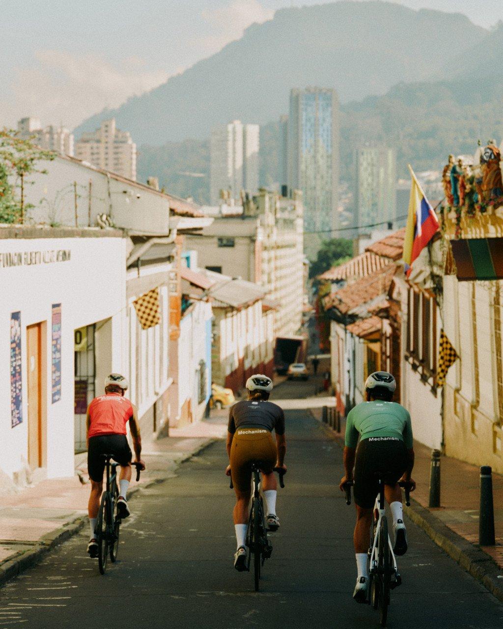 Three cyclists in Mechanism jerseys riding through colonial street with mountains and city skyline in background
