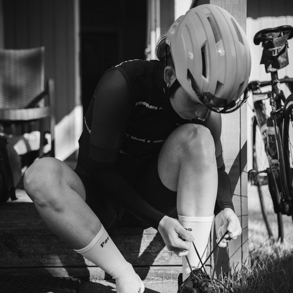 Model wearing black cycling jersey with white shorts and socks, tying shoes while wearing helmet