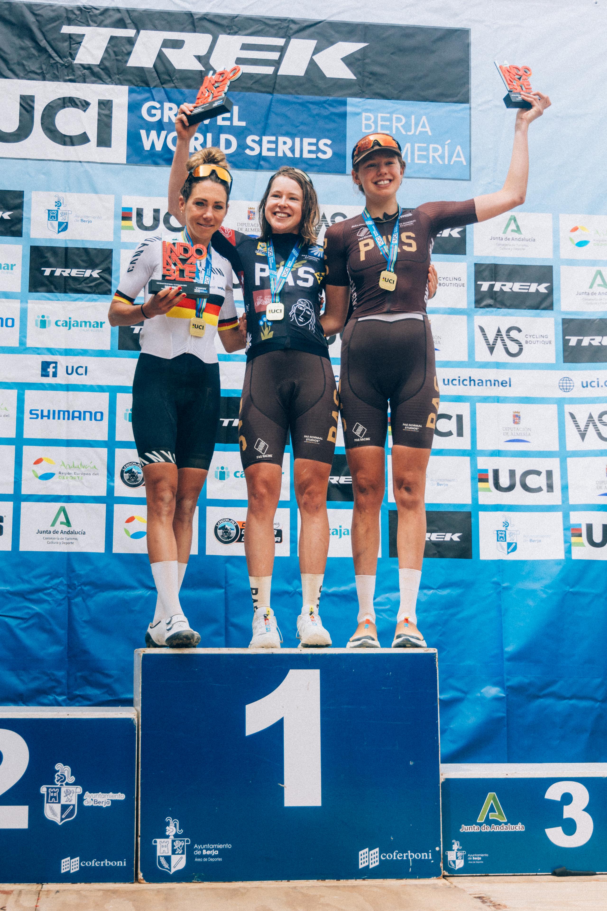 Three female cyclists on UCI race podium, winner wearing dark race kit with collection branding
